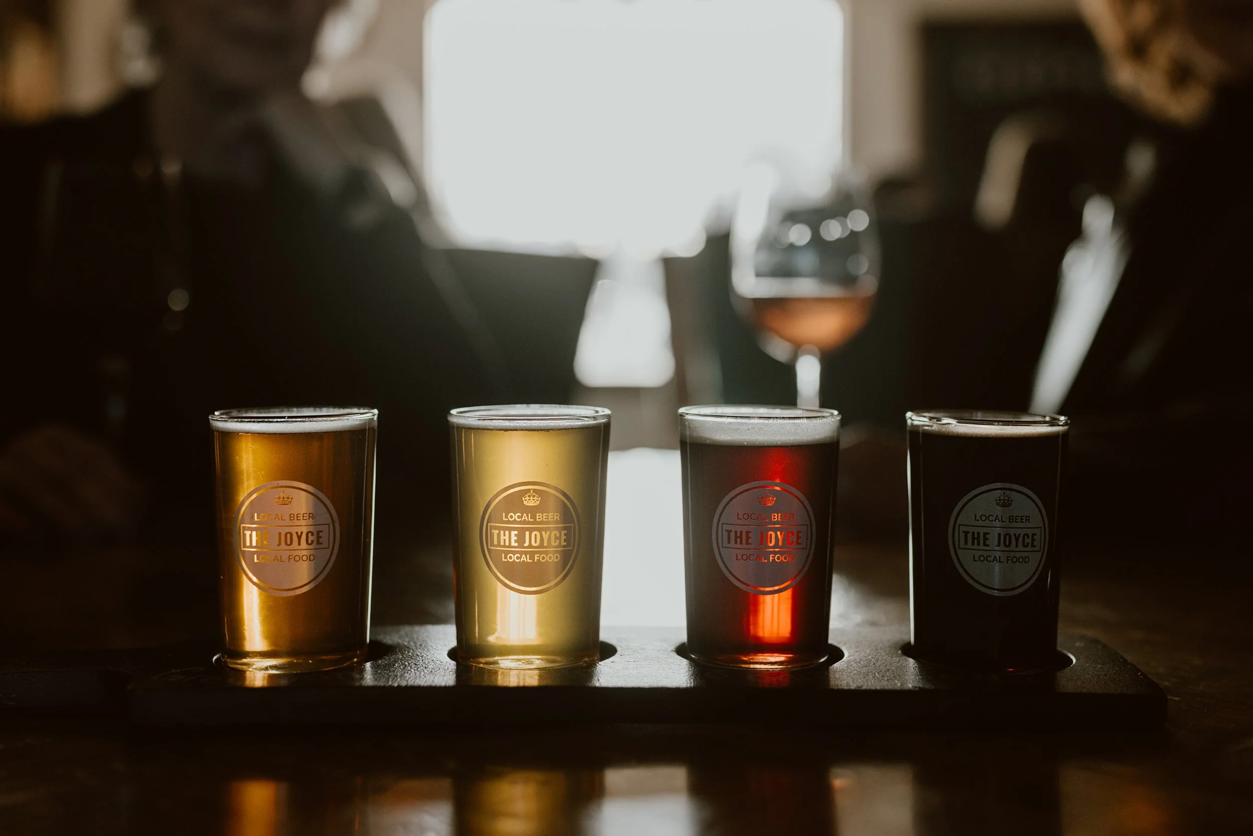 Four small glasses of different coloured craft beers on a dark beer flight paddle in the dimly lit Joyce Pub, with a person and a glass of rosé wine in the blurry background.