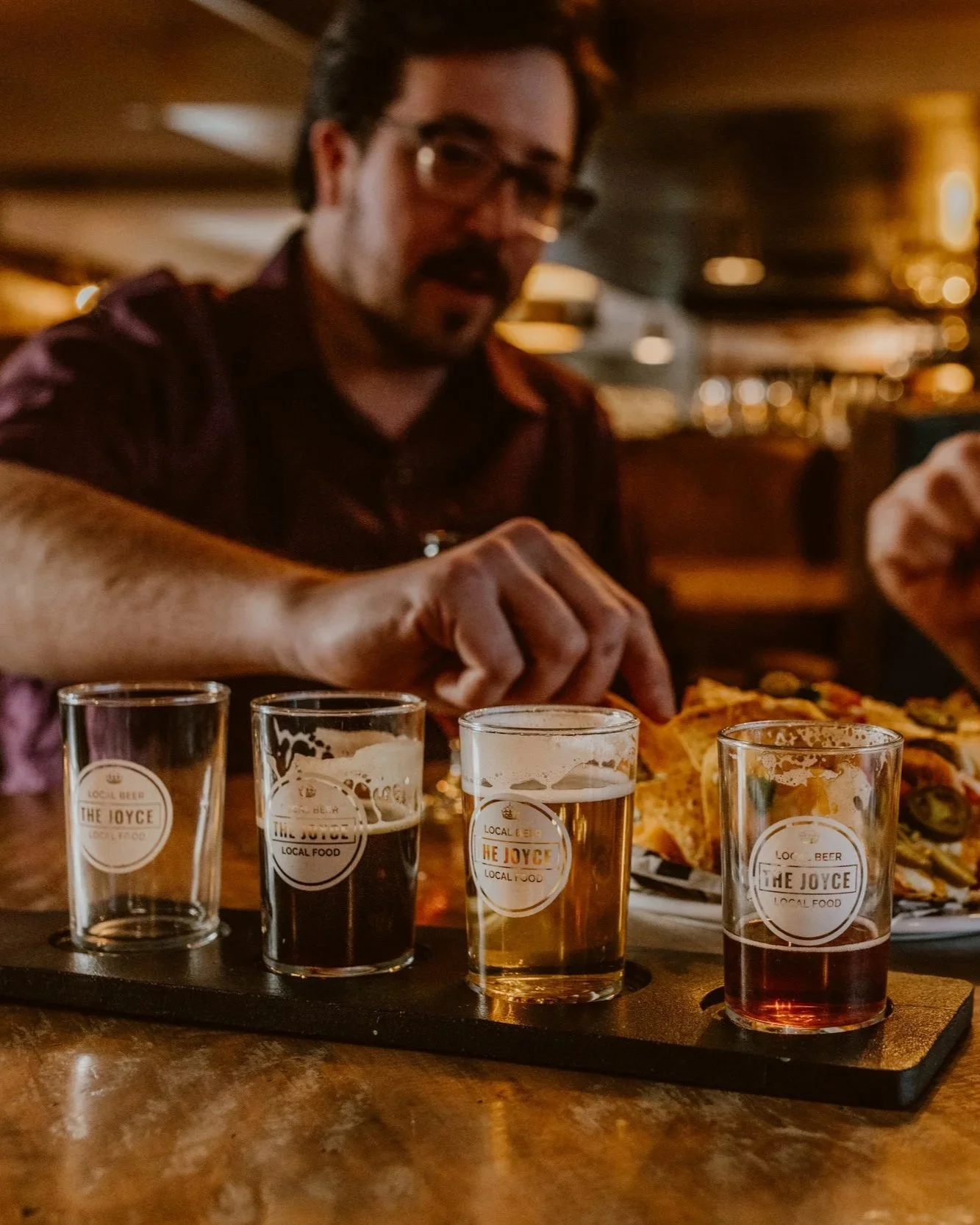 A man with glasses and a beard enjoying a craft beer flight at The Joyce Pub, with four tasting glasses filled with different types of craft beer on a tray, and a plate of nachos in the background.