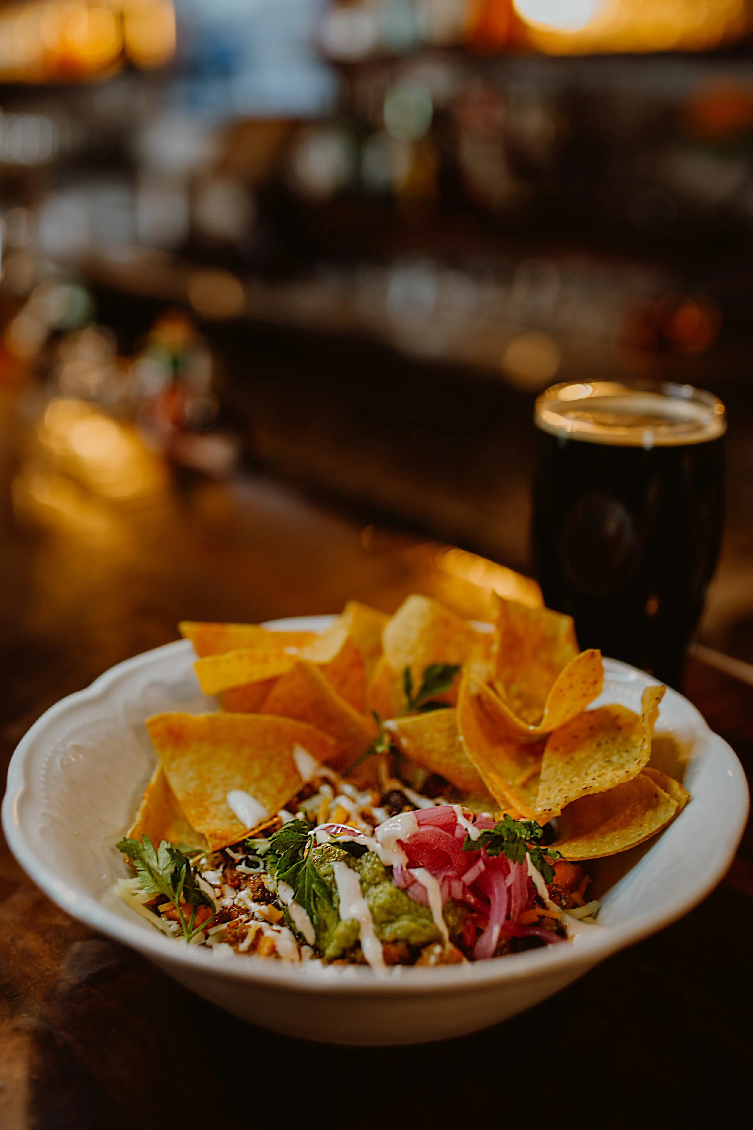 A bowl of nachos topped with salsa, cheese, guacamole, and cilantro, with a glass of dark craft beer in the background at The Joyce Pub.