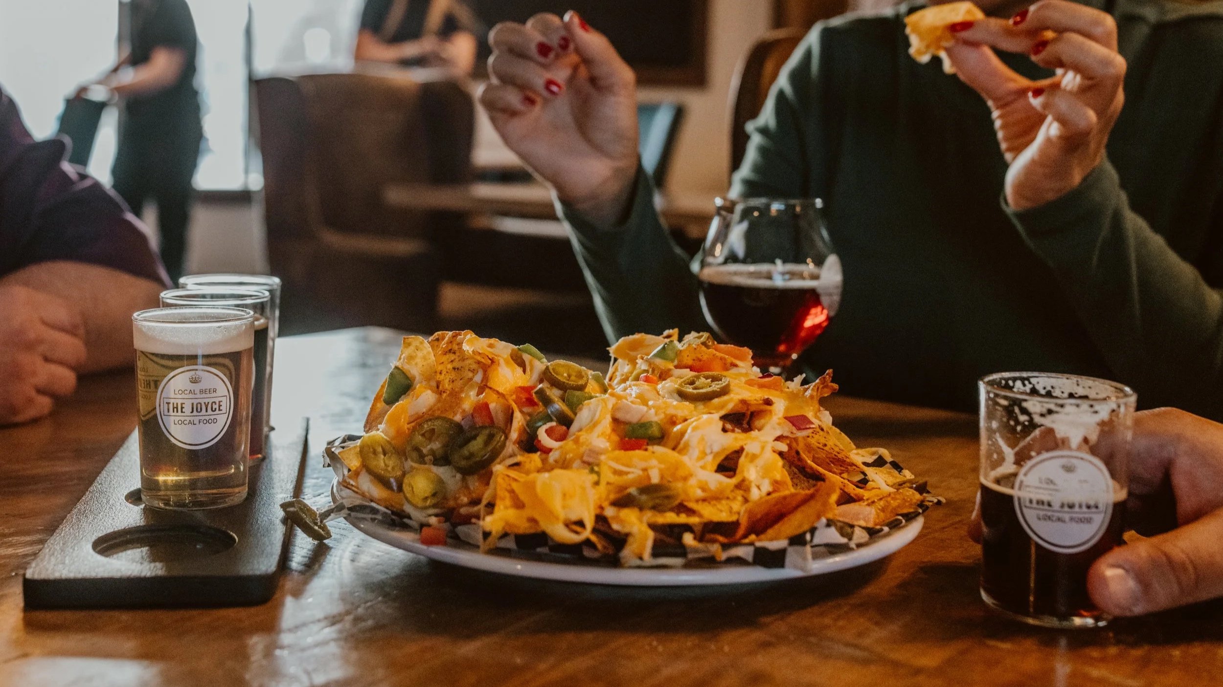 People sit around a table with a large plate of nachos topped with jalapeños and cheese, bottles of beer, and a glass of red wine, in the dimly lit dining area of The Joyce.