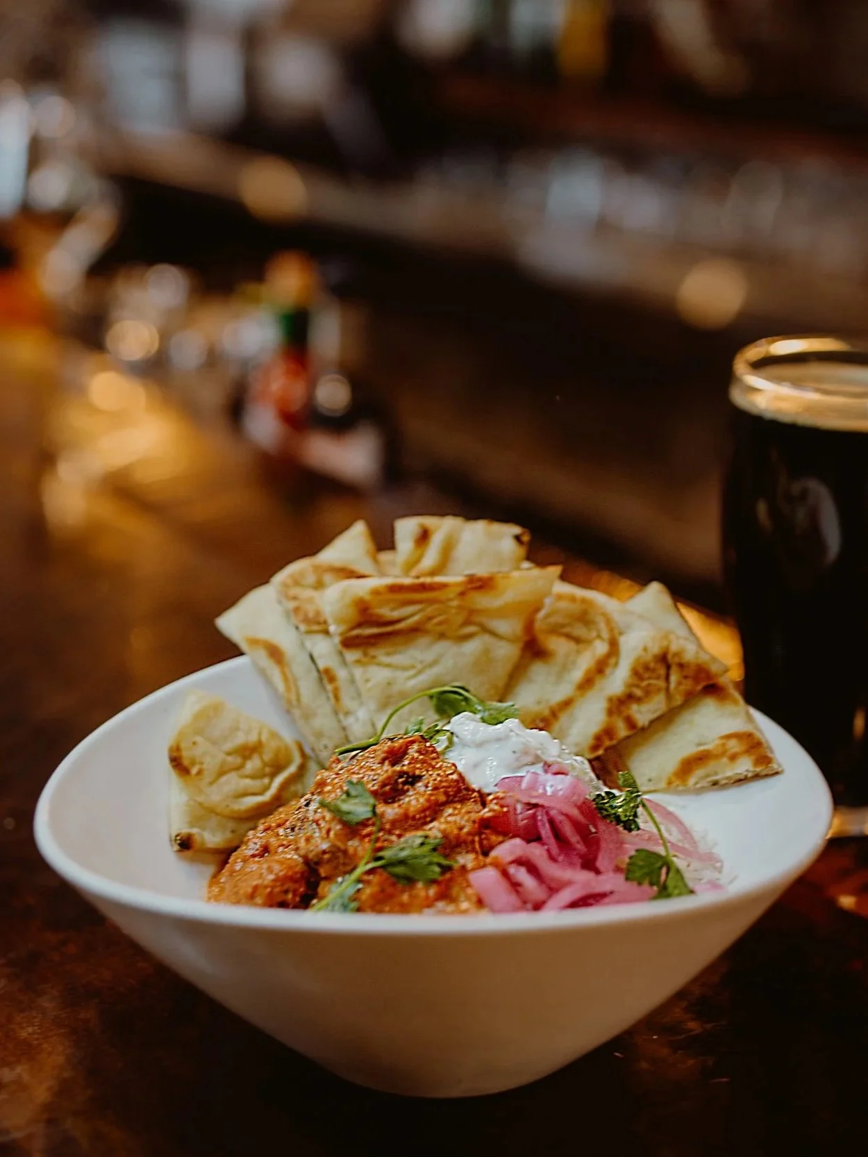A bowl of Indian food with naan bread, creamy chicken curry, pickled onions, and yogurt garnished with herbs, accompanied by a pint of dark craft beer served at The Joyce Pub.