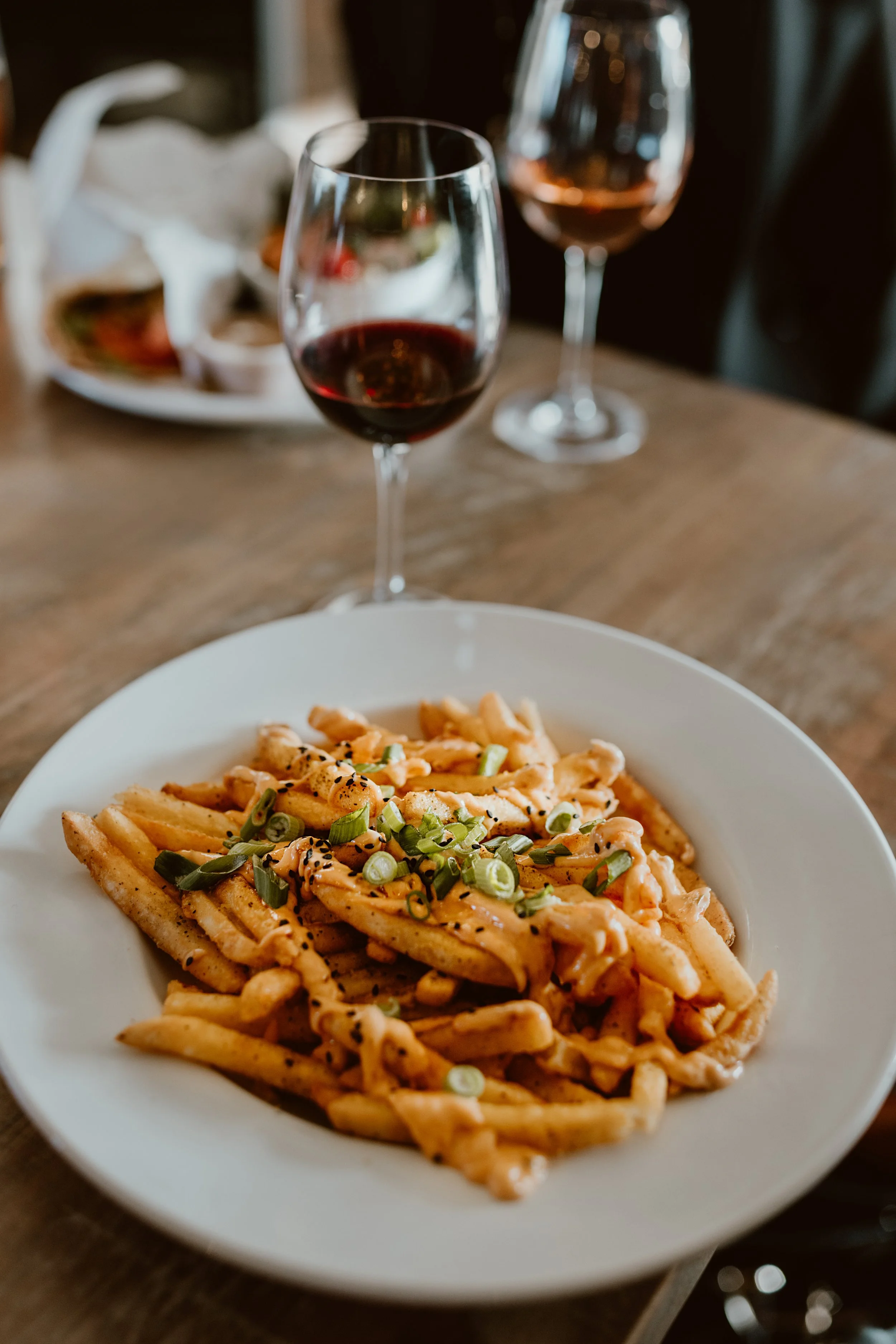 Plate of umami fries topped with chopped green onions, with two glasses of red wine in the background, served at The Joyce Pub.