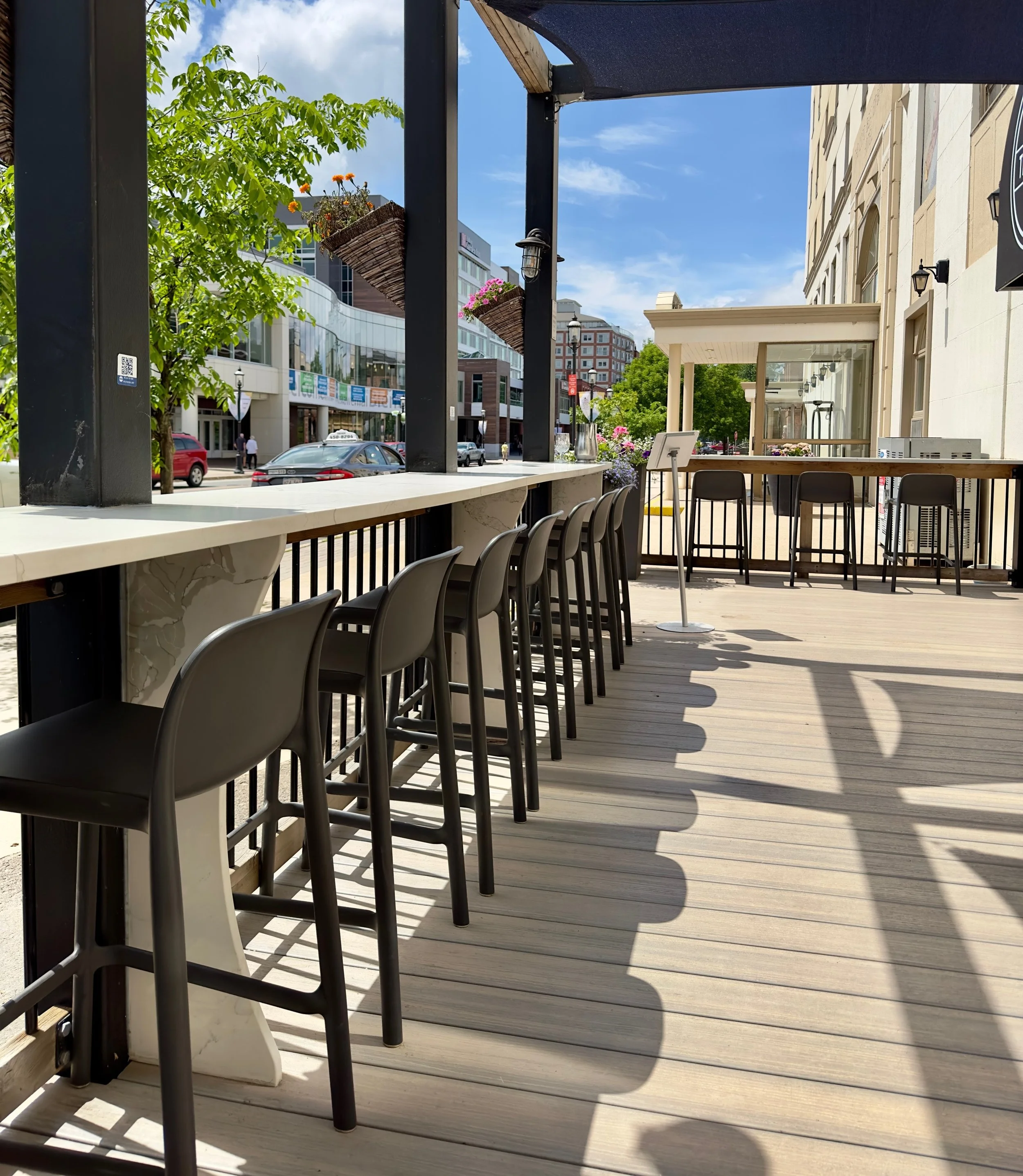 Empty outdoor patio of The Joyce Pub seating area with bar stools along a high counter, overlooking a city street with trees, cars, and modern buildings on a sunny day.