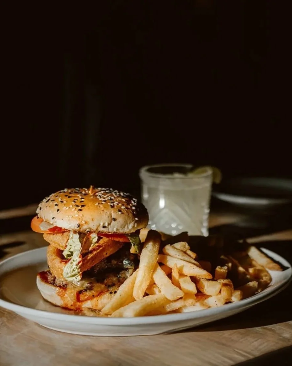 A cheeseburger with lettuce, tomato, pickles, and onions served with French fries on a white plate, with a cocktail in the background on a table at The Joyce Pub.