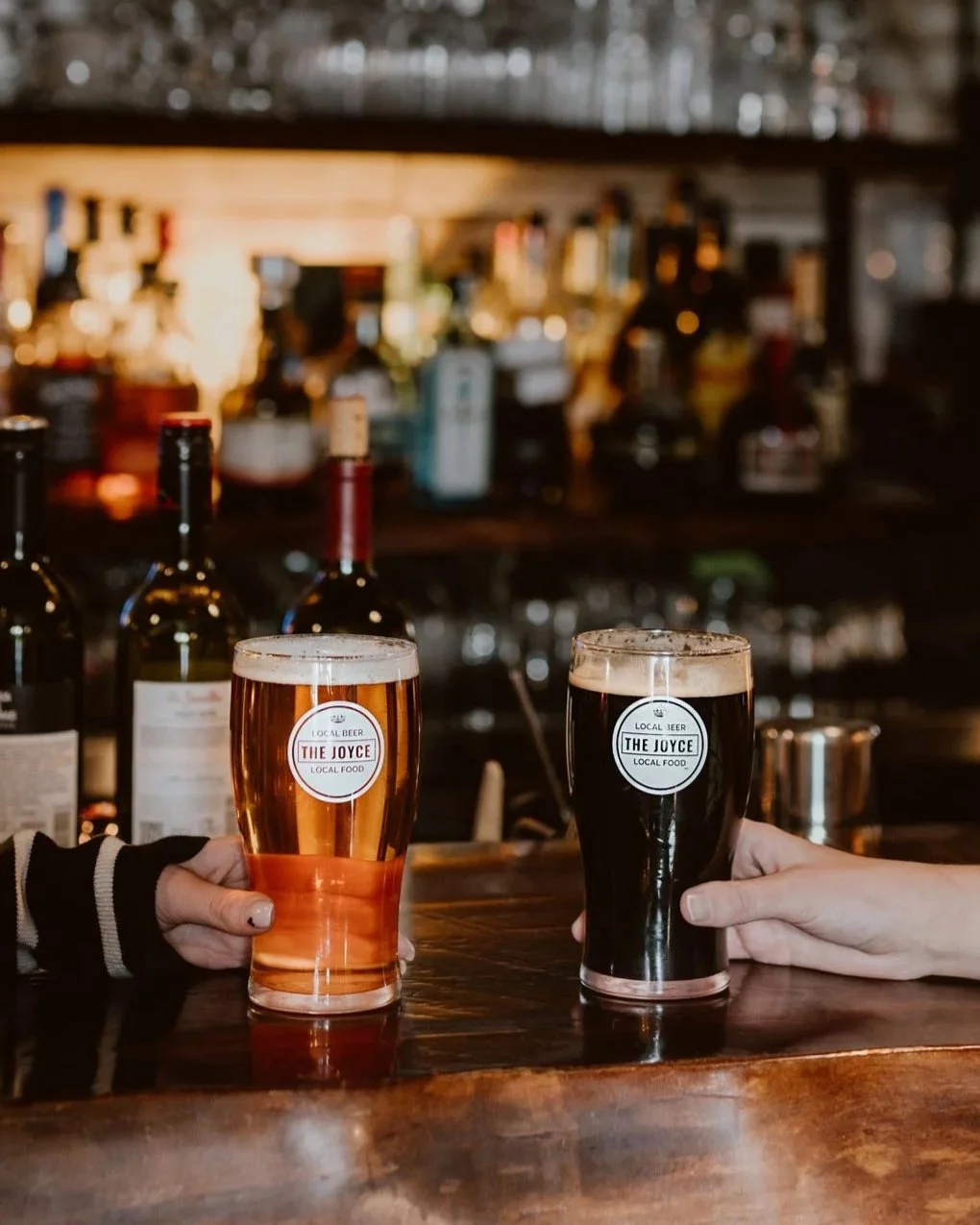 Two hands holding glasses of craft beer above the wooden bar top of The Joyce Pub with bottles in the background.