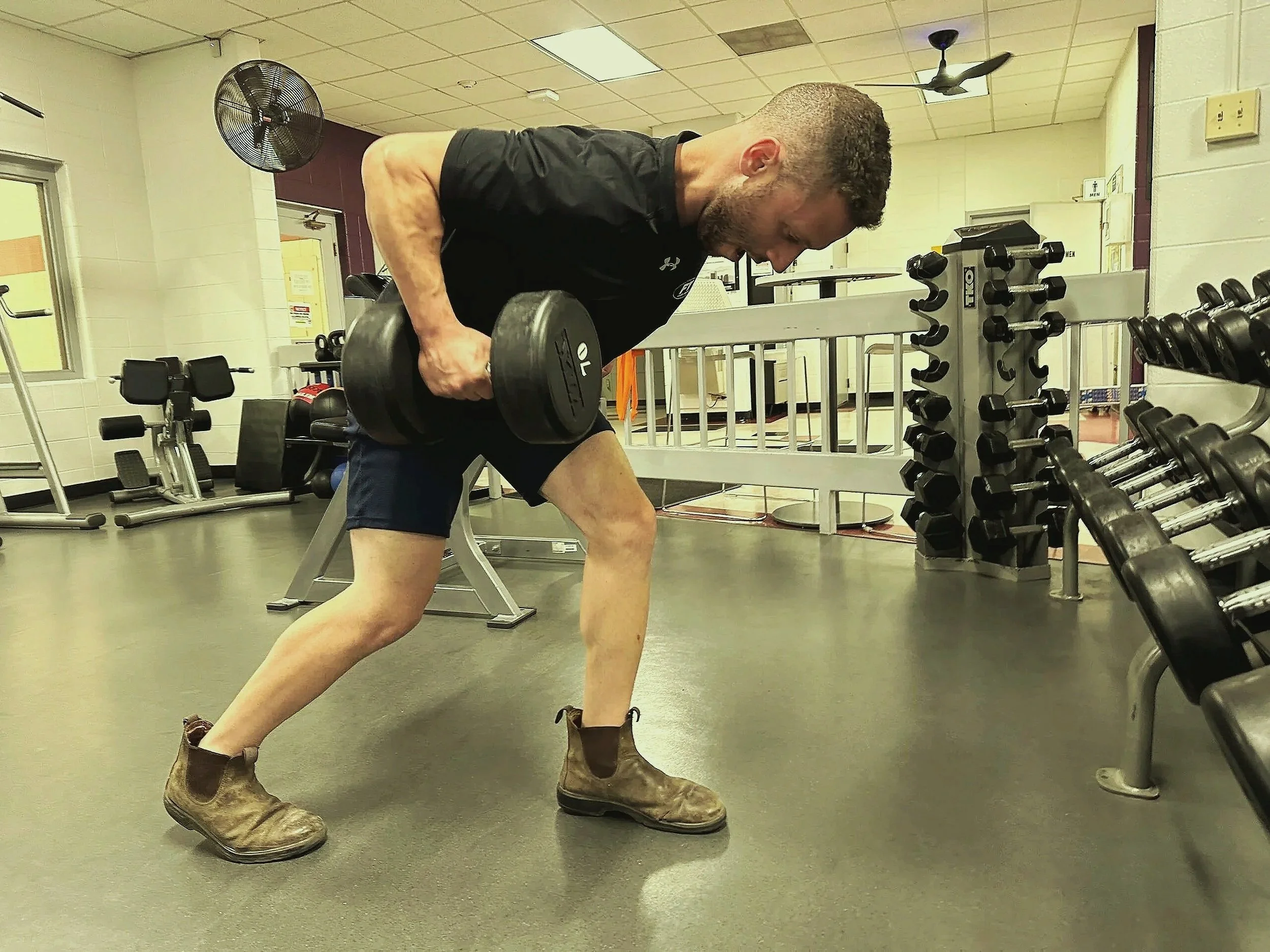 A man lifting a dumbbell in a gym, wearing a black T-shirt, dark shorts, and brown work boots, with gym equipment and weights in the background.