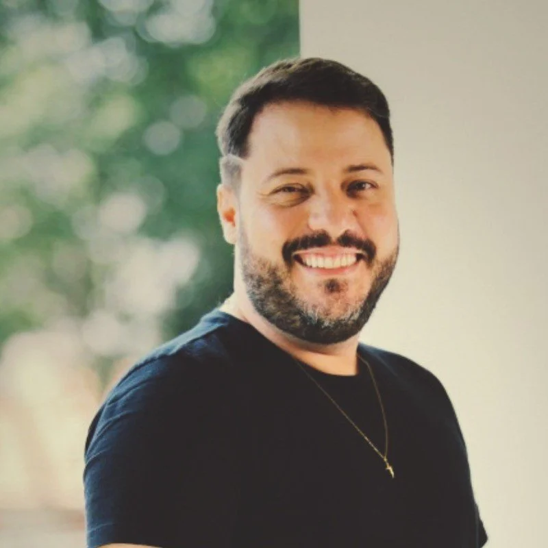 Homem sorrindo, barba e cabelo castanho, usando camiseta preta e um colar com cruz, ao fundo verde com bokeh