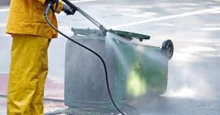 Person in yellow protective gear pressure washing a trash bin outdoors.