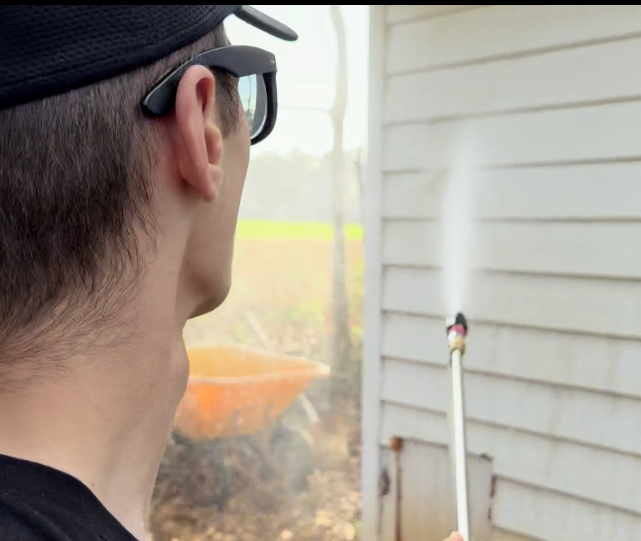 Person painting the exterior of a house with a paint roller, standing on a ladder. The house is white with blue shutters, and there are tall bushes nearby.