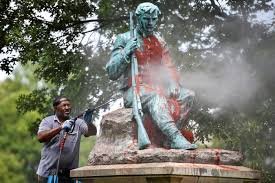 A person cleaning a large statue of a seated man outdoors surrounded by trees, with mist or spray around the statue.