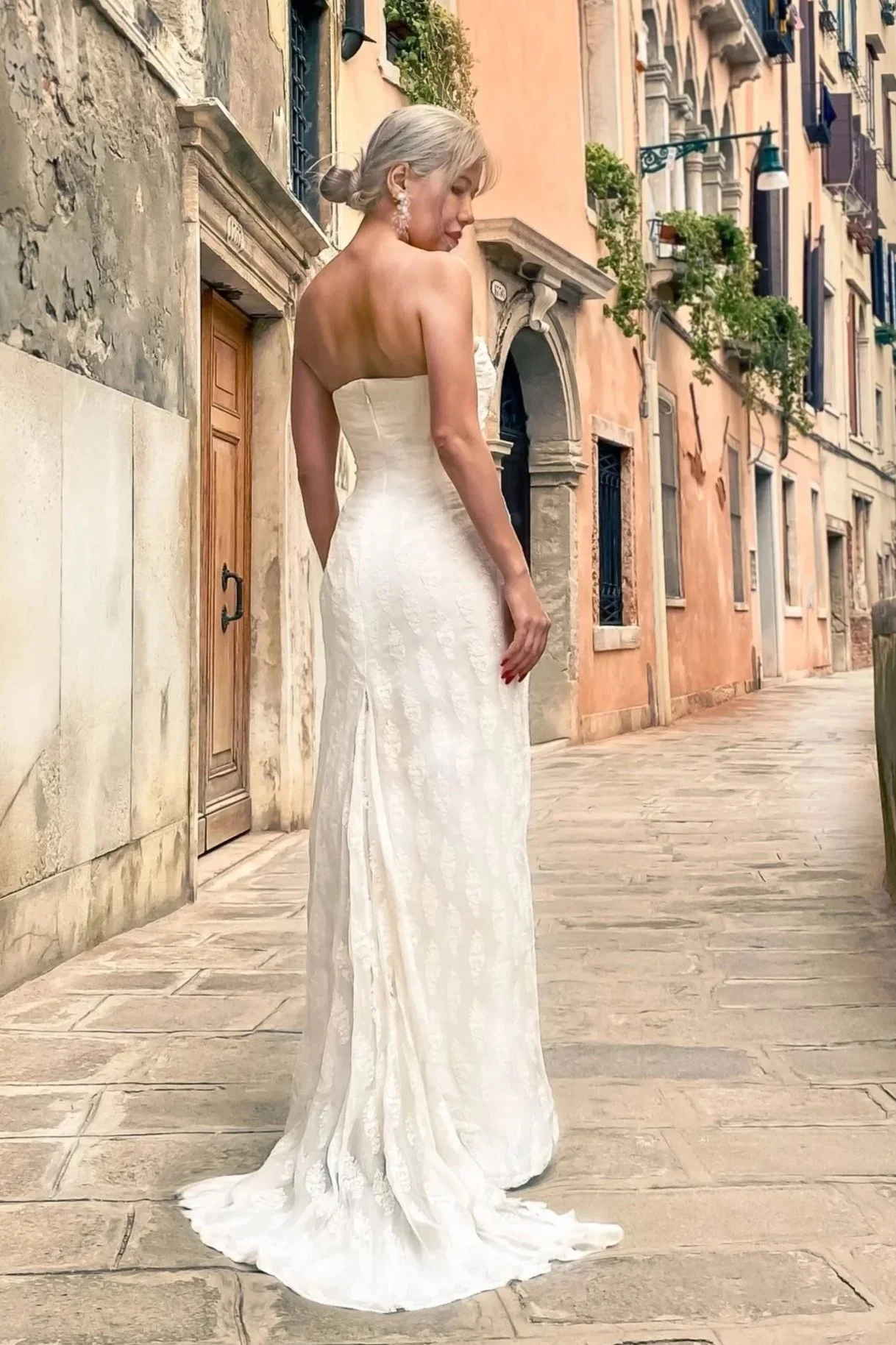 A woman in a strapless white wedding dress stands on a European-style street with old buildings and potted plants, facing sideways with her head slightly turned down.