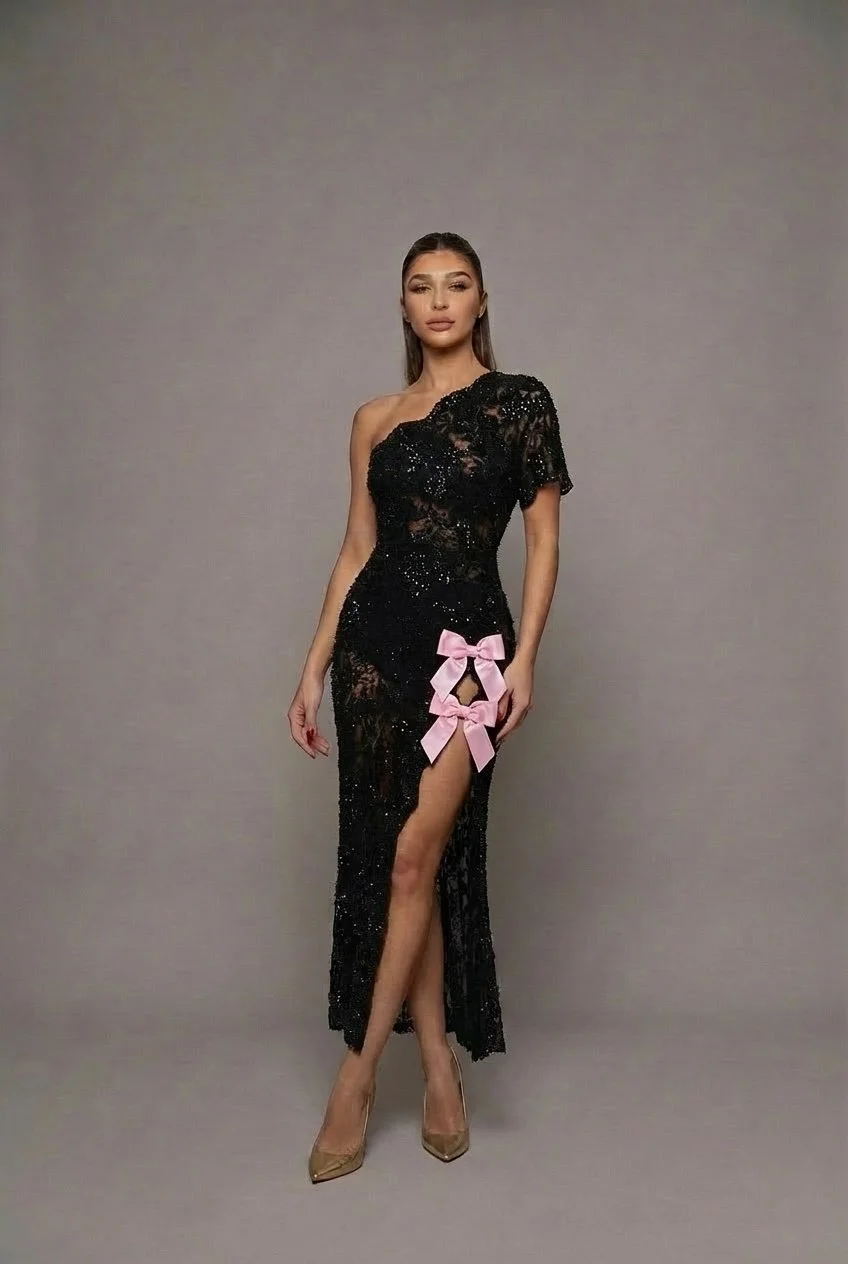 Black one-shoulder sequined lace dress with a fitted, body-contouring silhouette.
Asymmetrical neckline with one short lace sleeve. Slim midi column shape with a high side slit.
Two pale pink satin bows accent the slit, adding contrast to the dark, s
