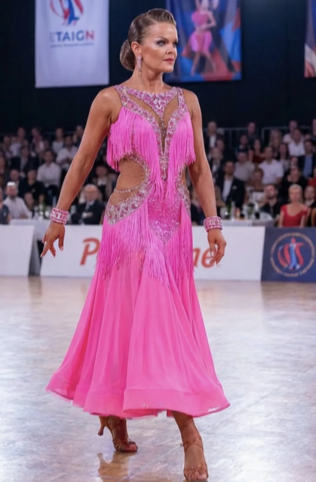Woman in a bright pink ballroom dance dress with fringe and cutouts, walking on a dance floor during a competition, with an audience in the background.