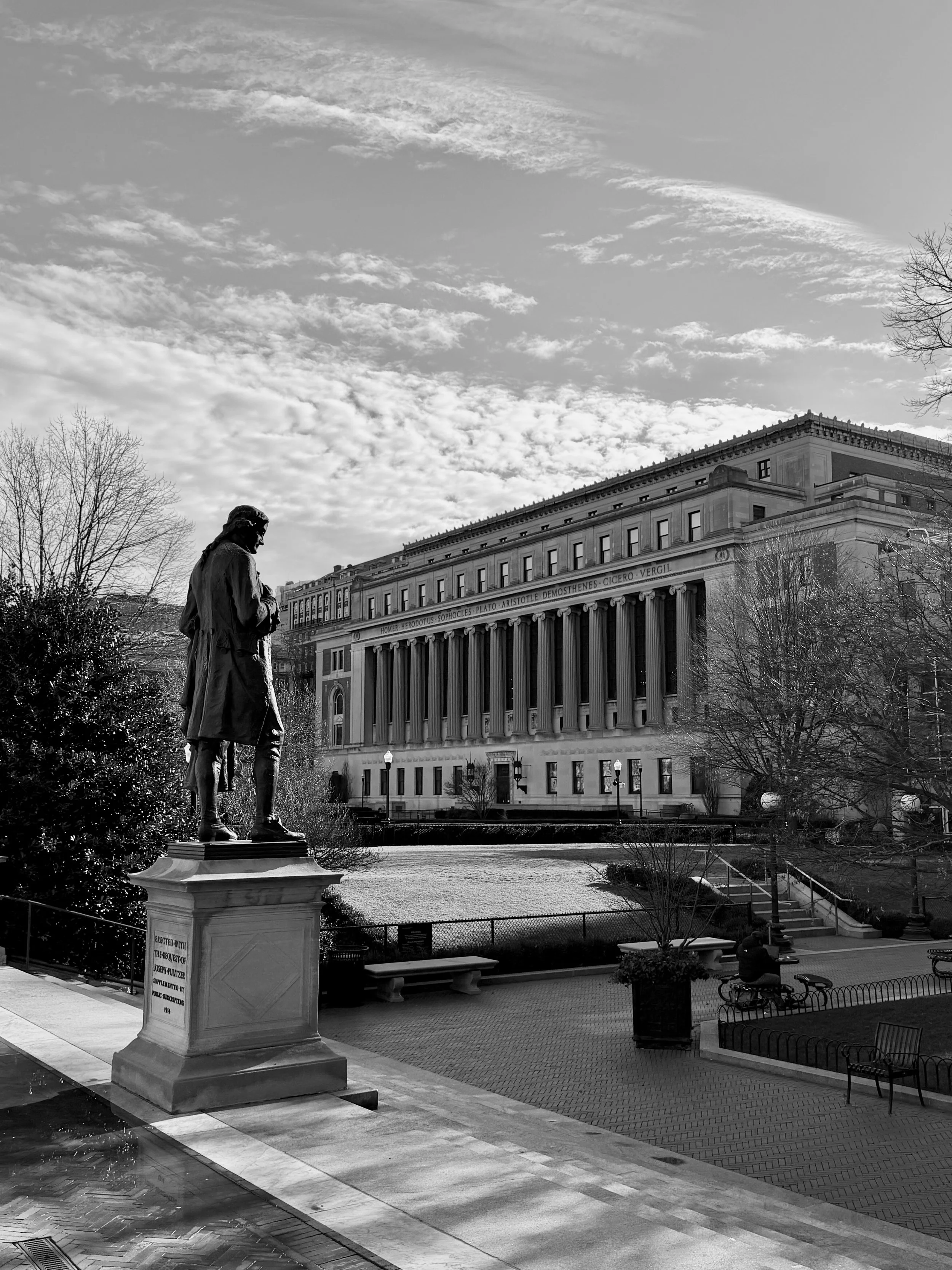Black and white photo of a park with a statue of a man on a pedestal, trees, benches, and a neoclassical government building in the background, under a partly cloudy sky.