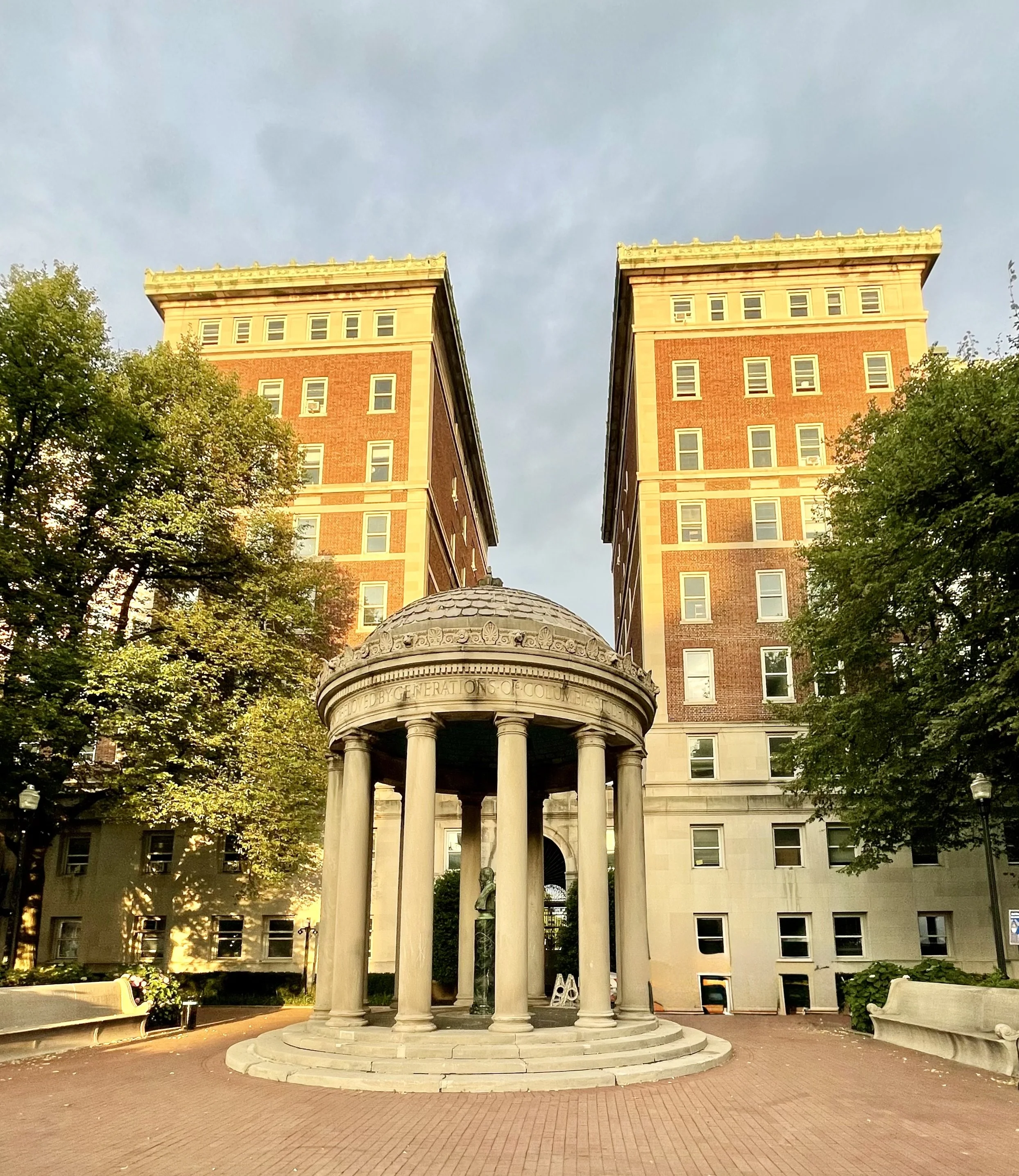 A park with trees, benches, and a small round stone pavilion with Roman columns and a dome, with tall brick and stone buildings in the background under a cloudy sky.