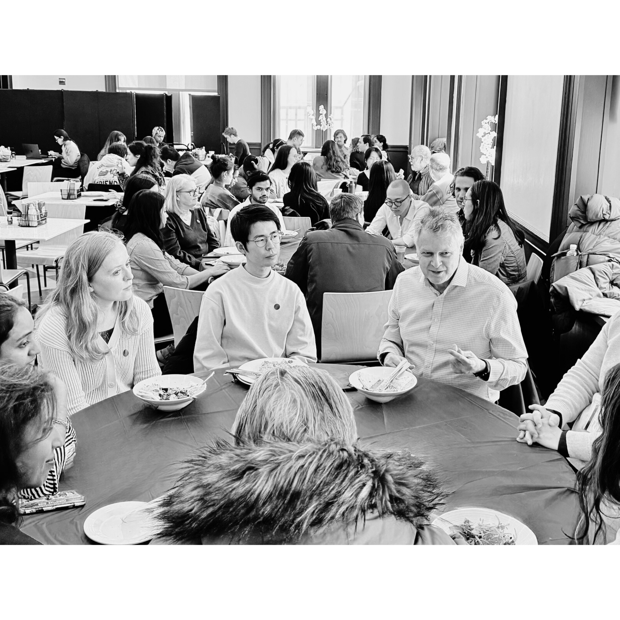 People gathered around a table at a social event or meeting in a well-lit room with large windows.