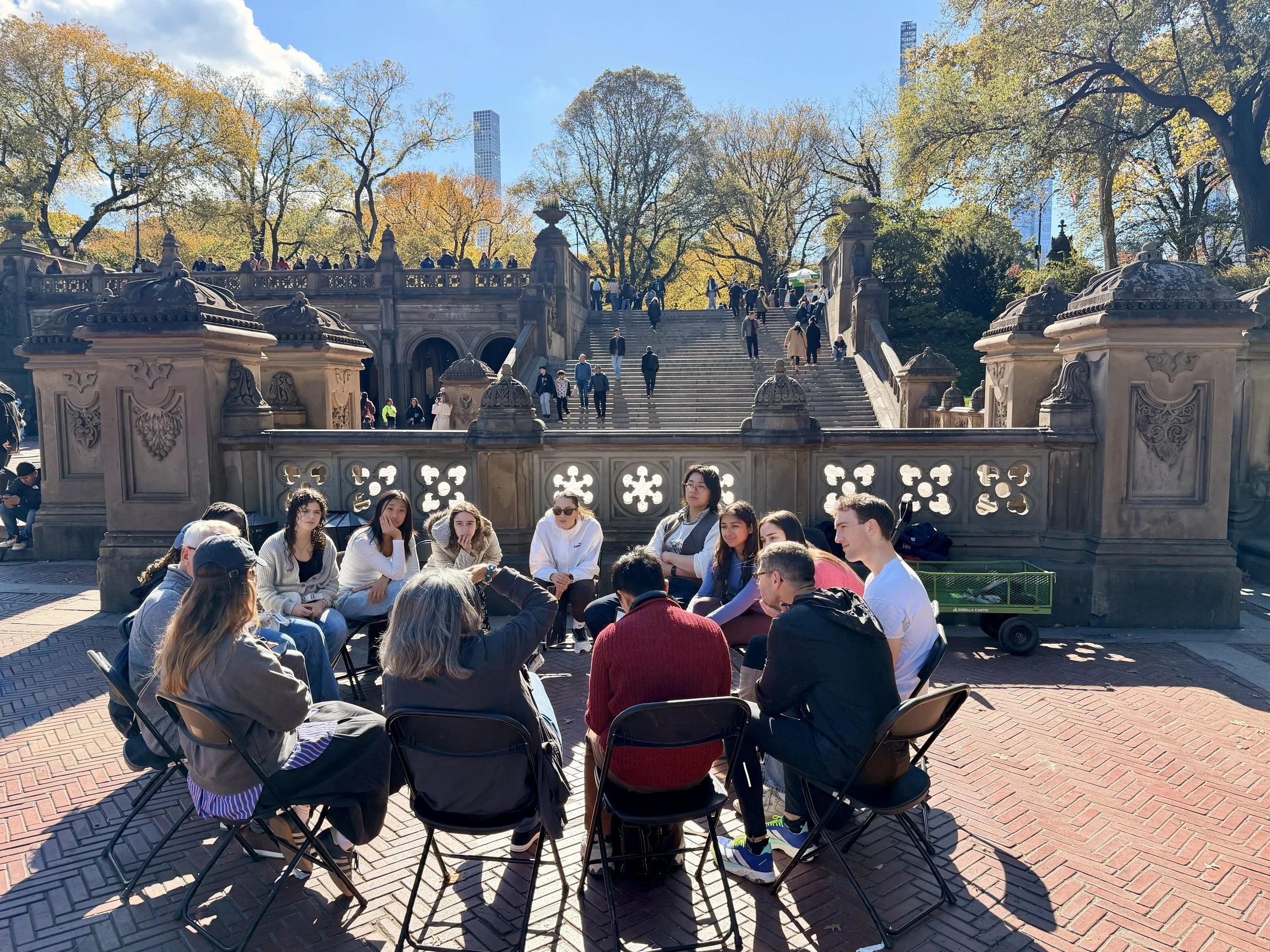 A group of people sitting in a circle outdoors on a sunny day, near a stone staircase and decorated fence, with trees and skyscrapers in the background.