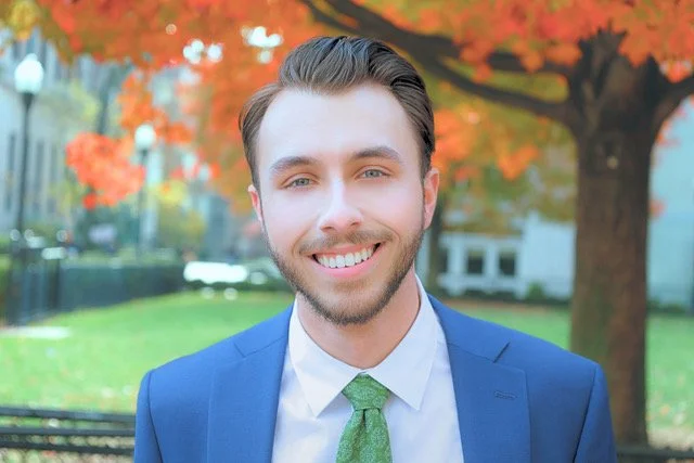 A young man with light brown hair and a beard, wearing a blue suit and green tie, smiling outdoors in front of trees with orange autumn leaves.
