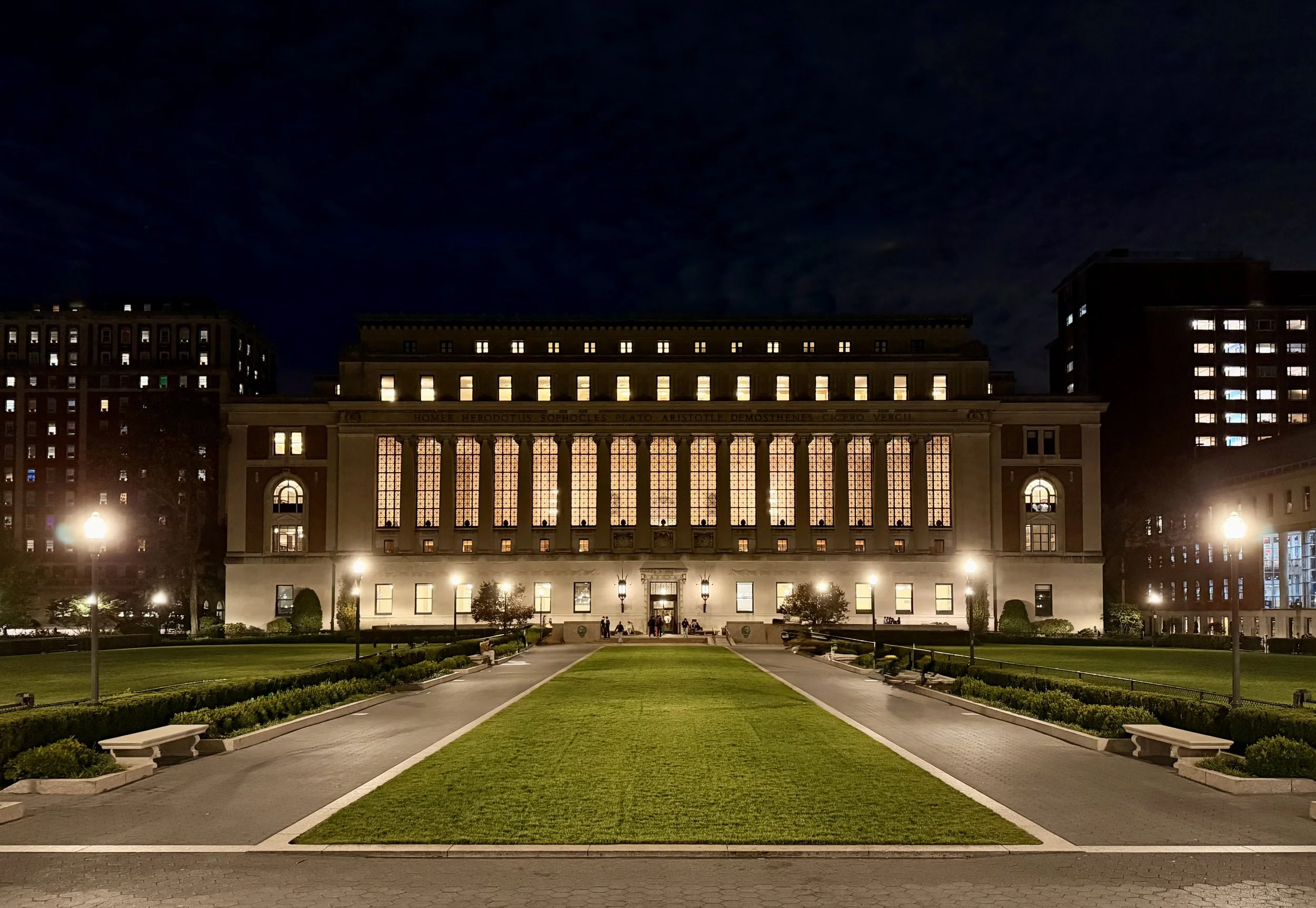 Nighttime view of a lit-up historic building with large stained-glass windows, surrounded by a well-maintained park with benches, walkway, and street lamps.