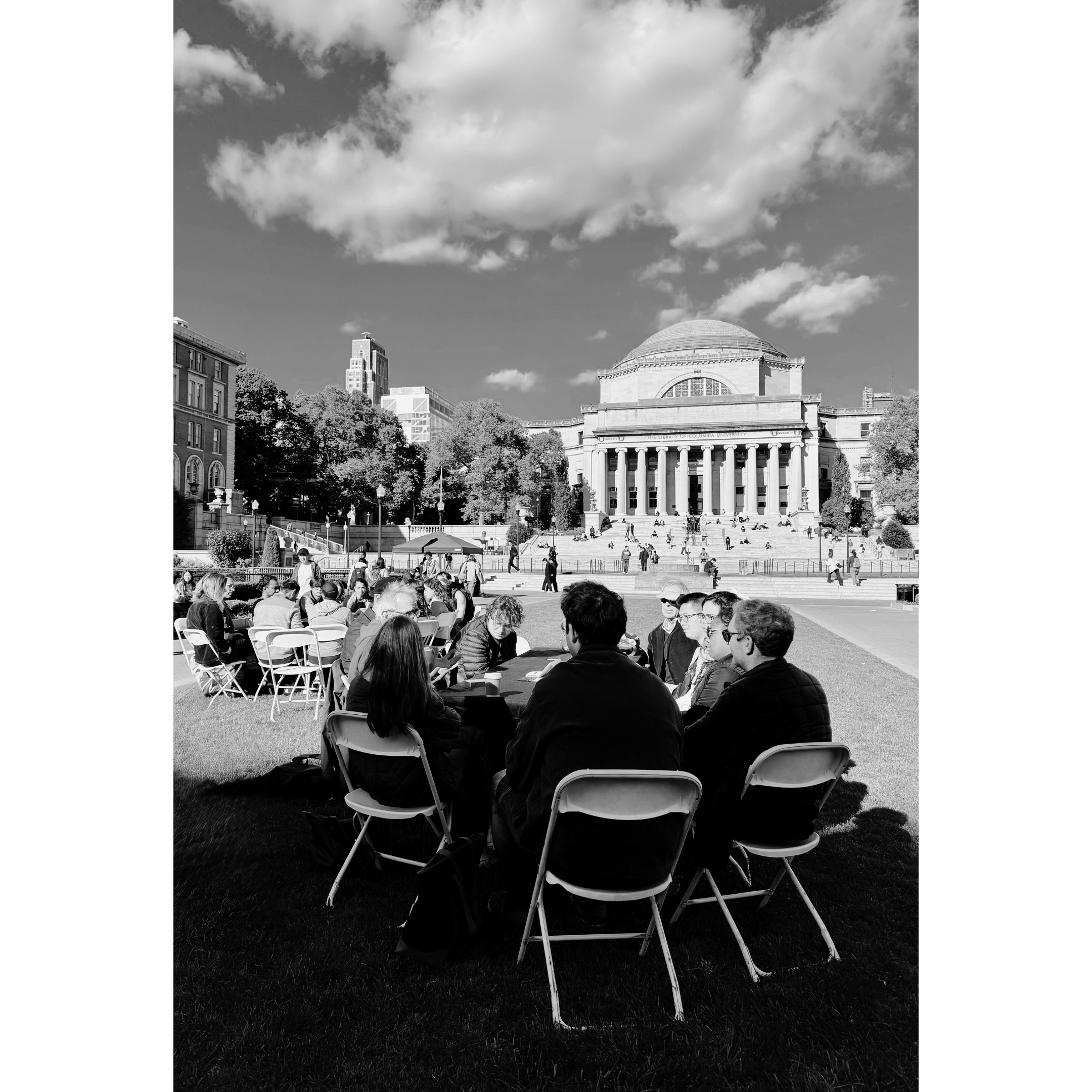 A group of people sitting in chairs on a lawn, with a large neoclassical building with columns and a dome in the background, under a partly cloudy sky.