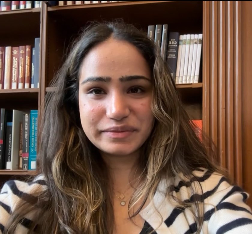 A young woman with long, wavy brown hair taking a selfie in front of a bookshelf filled with books.