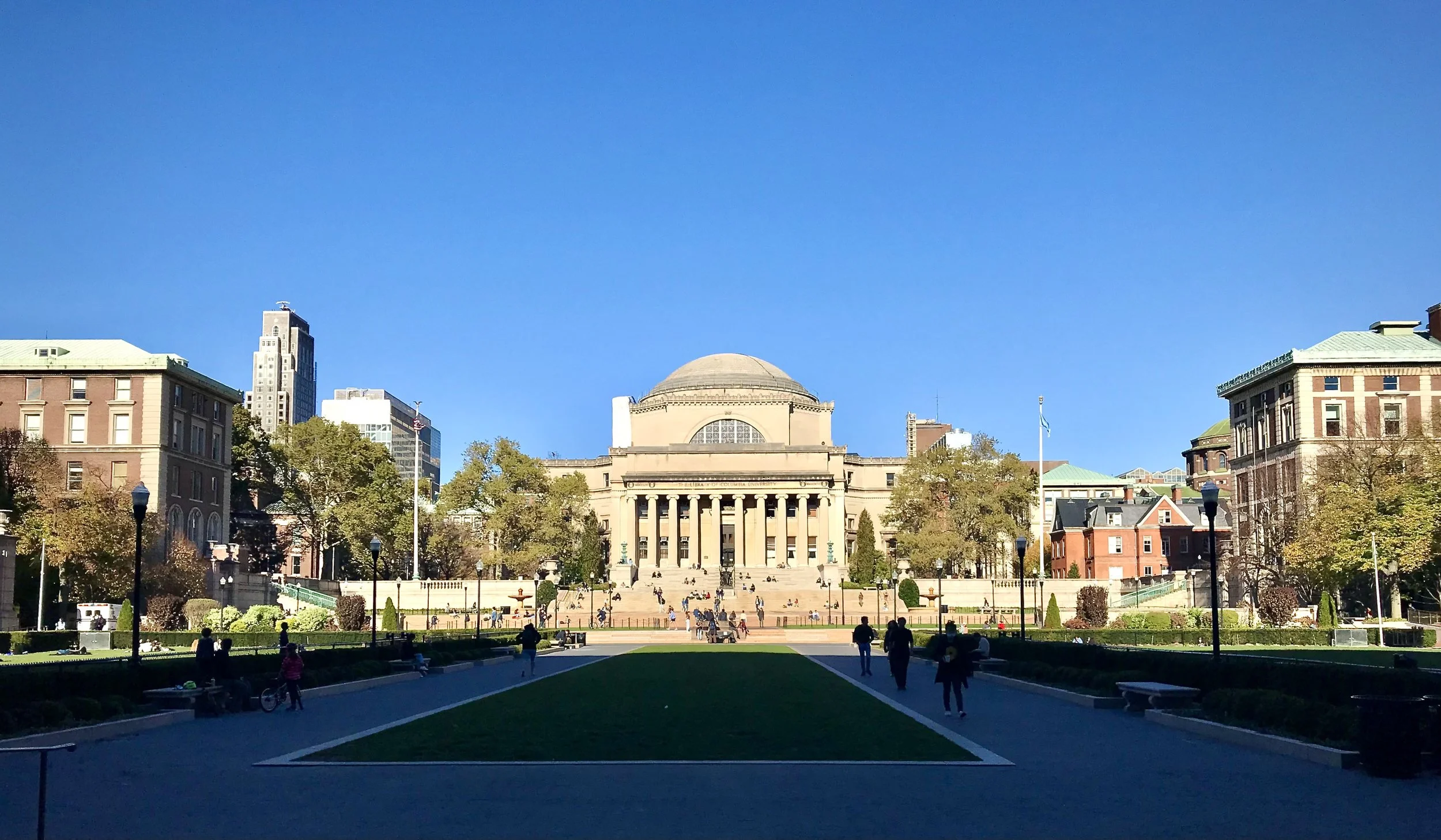 A grand neoclassical building with a dome, columns, and steps, surrounded by trees, in front of a well-maintained park with people walking and sitting, under a clear blue sky.