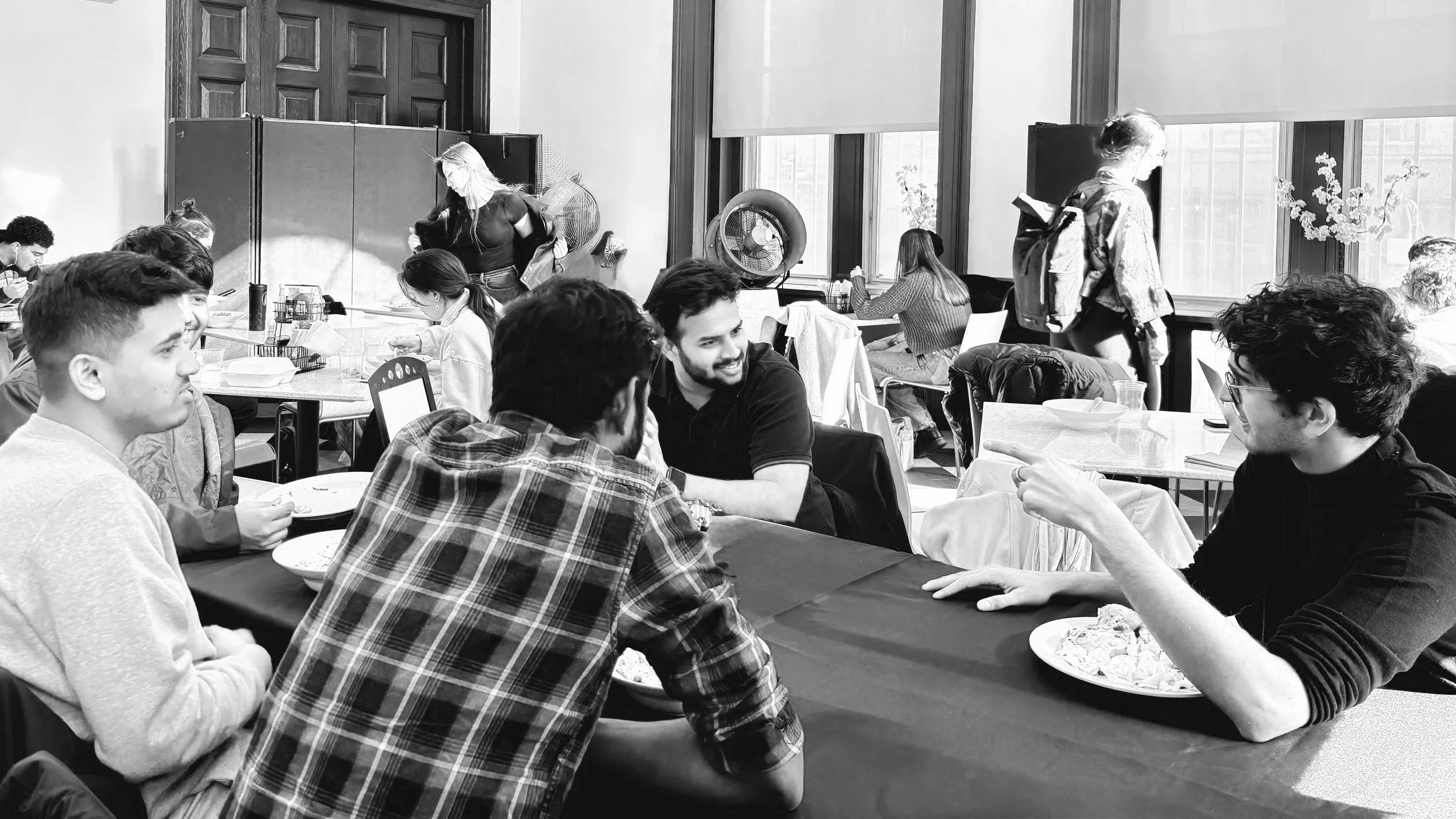 A group of people sitting around a dining table engaged in conversation in a well-lit room with large windows. Some individuals are eating, chatting, or listening, with others in the background.