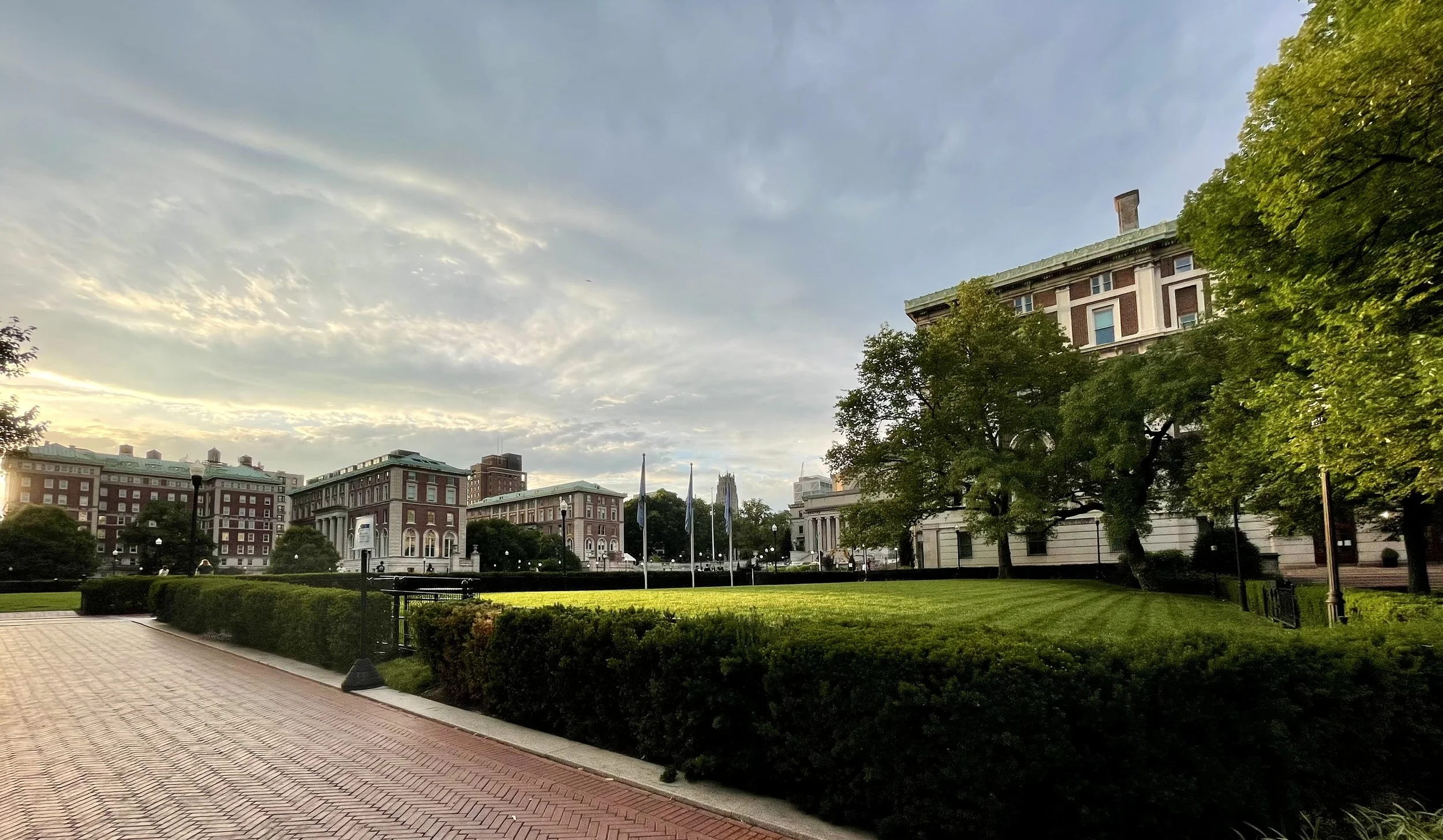 City park with green grass, trees, and historic brick buildings, under a partly cloudy sky at sunset.
