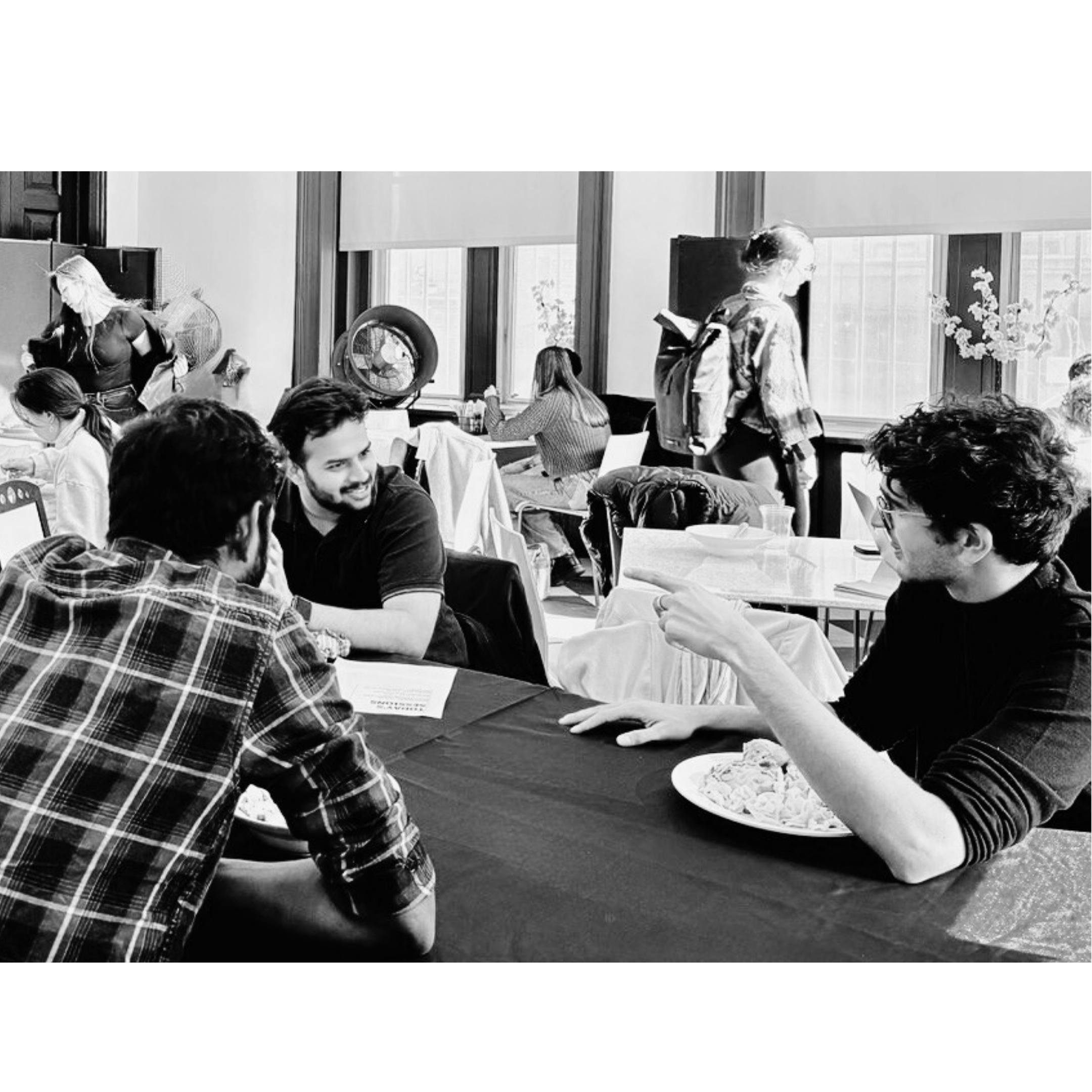 Group of young people talking and socializing at a table in a casual indoor setting, with some women working or sitting near windows in the background.