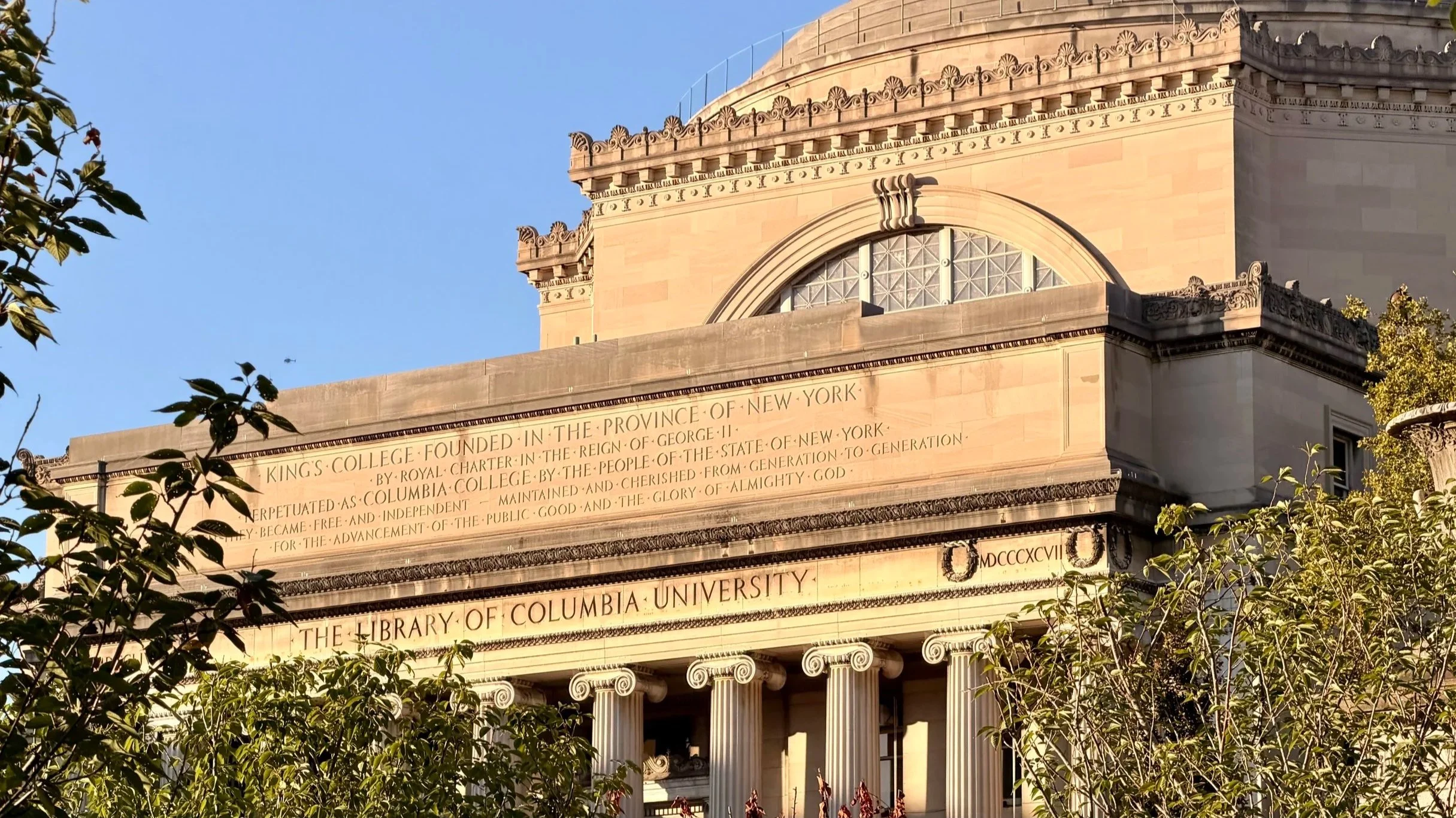 The library of Columbia University, a classical building with columns and engraved inscriptions, surrounded by trees and foliage, under a clear blue sky.