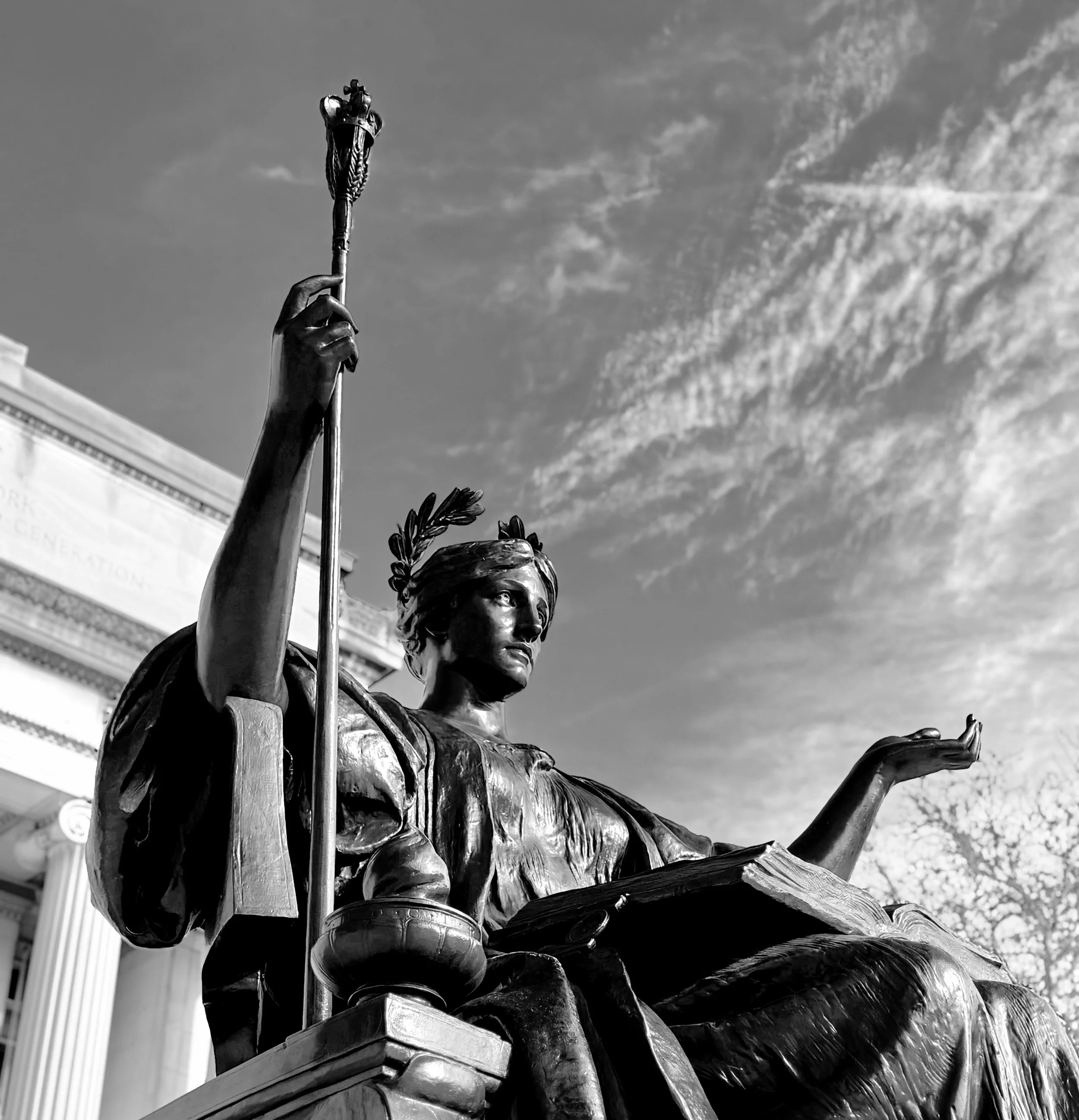 Black and white photo of a bronze statue of a seated woman holding a staff, with a building in the background and sky with wispy clouds.