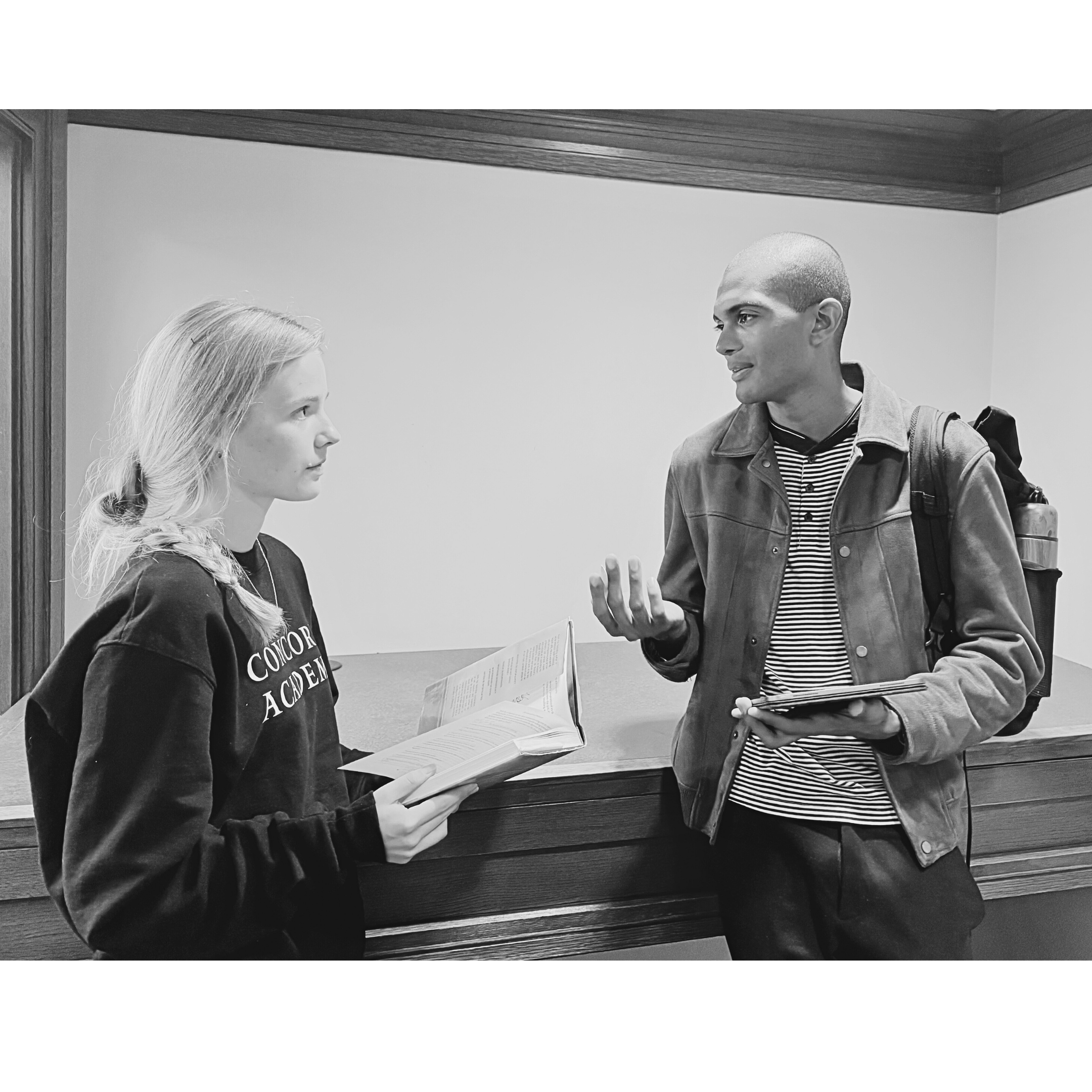 Two young adults, a woman and a man, engaged in conversation at a reception desk. The woman holds an open book, while the man has a notebook or tablet. The setting appears to be an indoor institutional environment.