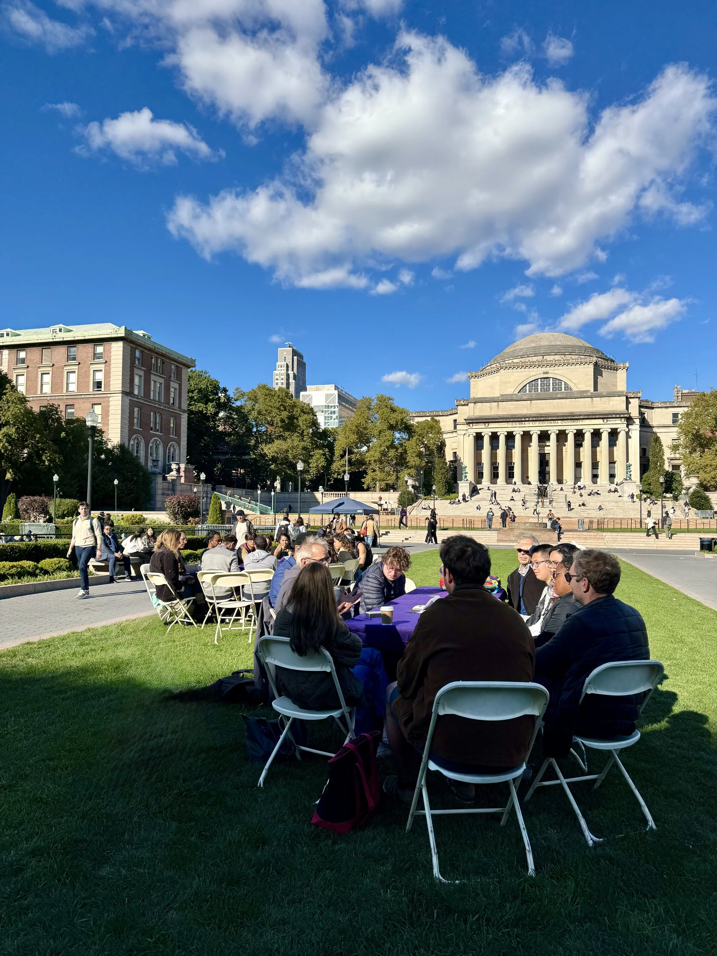 Group of people sitting at tables on a grassy lawn in front of a large neoclassical building with columns, surrounded by trees and city buildings, on a sunny day with blue sky and clouds.