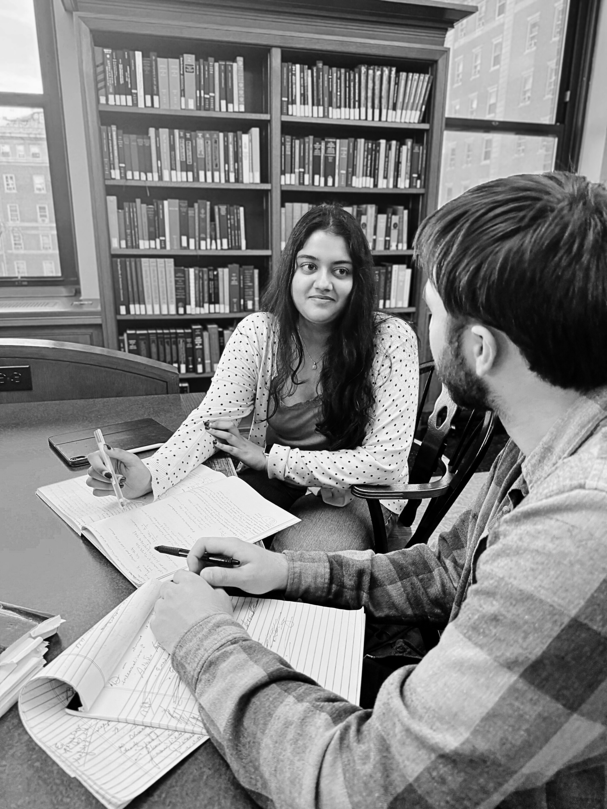 Two students, a woman with long dark hair and a man with short hair and a beard, are sitting at a table in a library, engaged in a discussion with open notebooks and pens. Behind them, there are bookshelves filled with books and a window showing buildings outside.