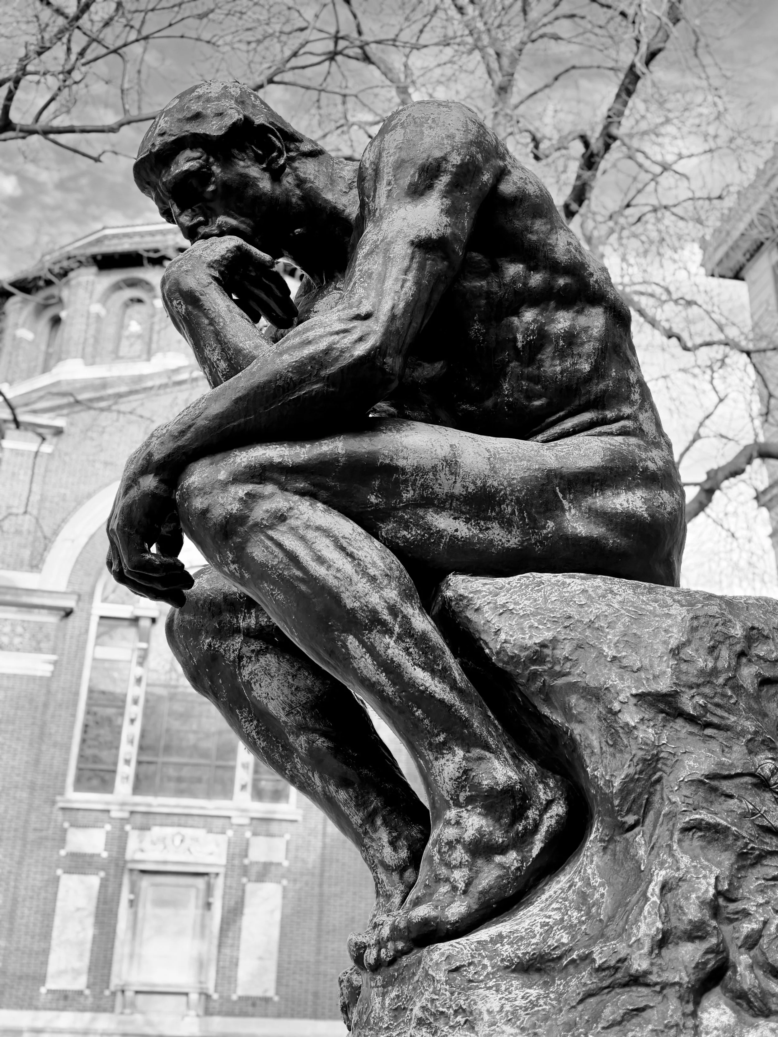 Close-up of the Thinker sculpture by Auguste Rodin, showing a contemplative man sitting on a rock with his chin resting on his hand, outdoors with a building and leafless trees in the background.