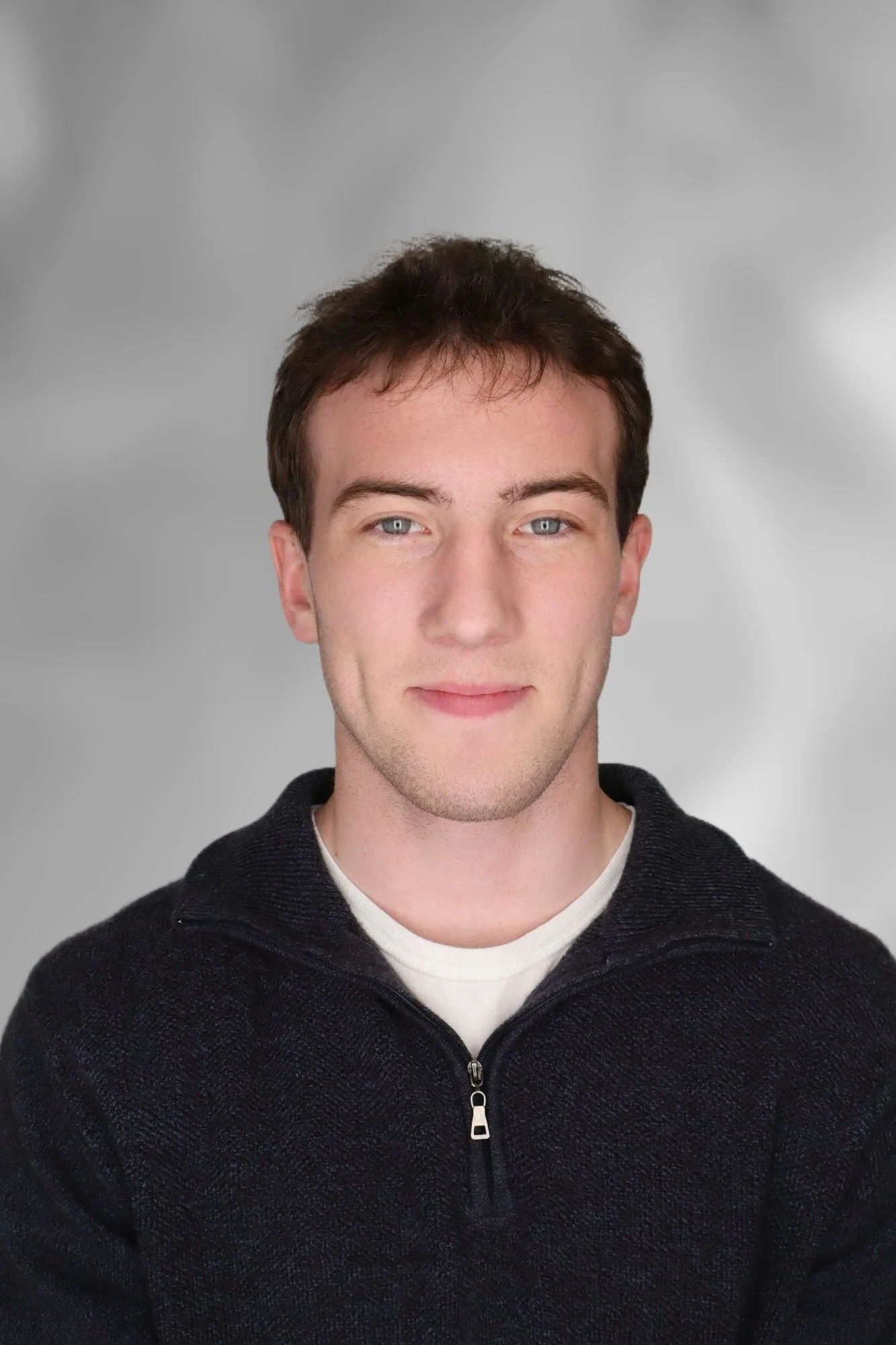 A young man with short, dark hair, light skin, and blue eyes, wearing a black zip-up sweater over a white shirt, looking directly at the camera with a light smile, against a plain gray background.