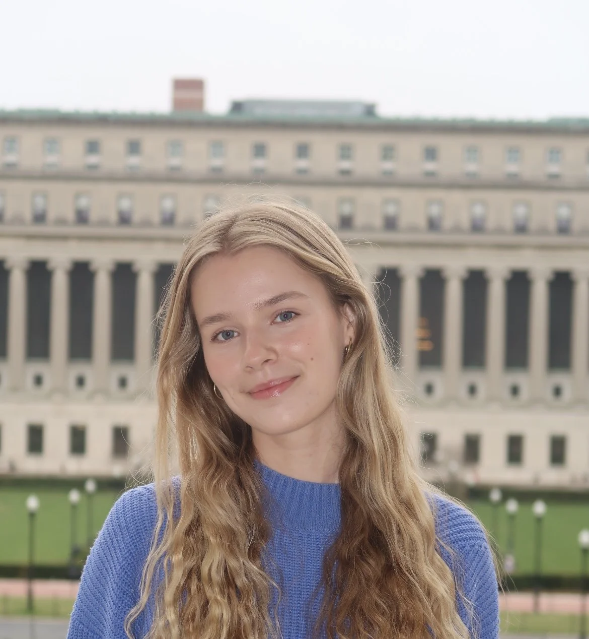 A young woman with long wavy blonde hair, wearing a blue sweater, standing outdoors in front of a large building with columns and windows.