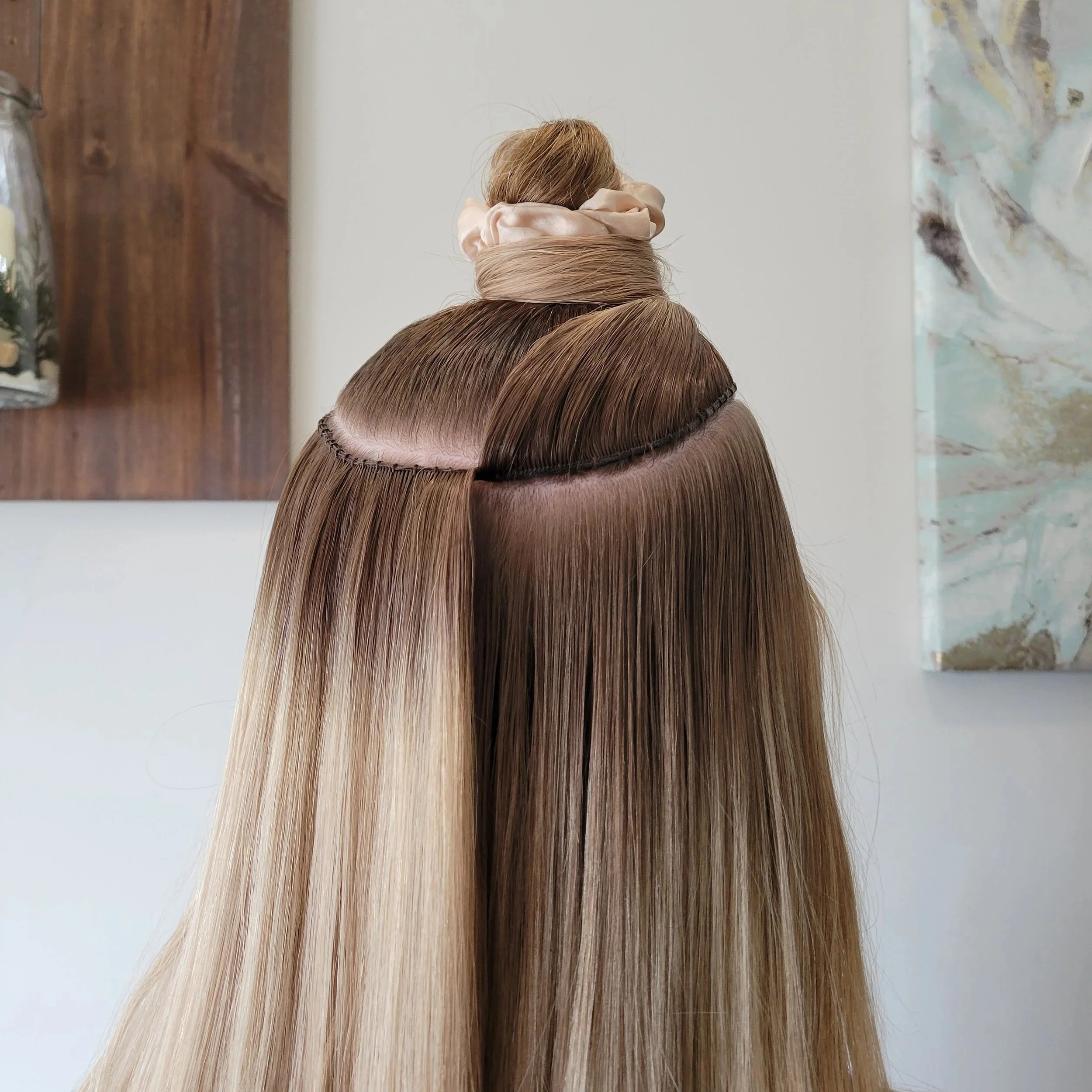 Close-up of a hair styling station with long, straight, ombré hair and a sewing weft attached to a section of hair, with a hair bun in the background.