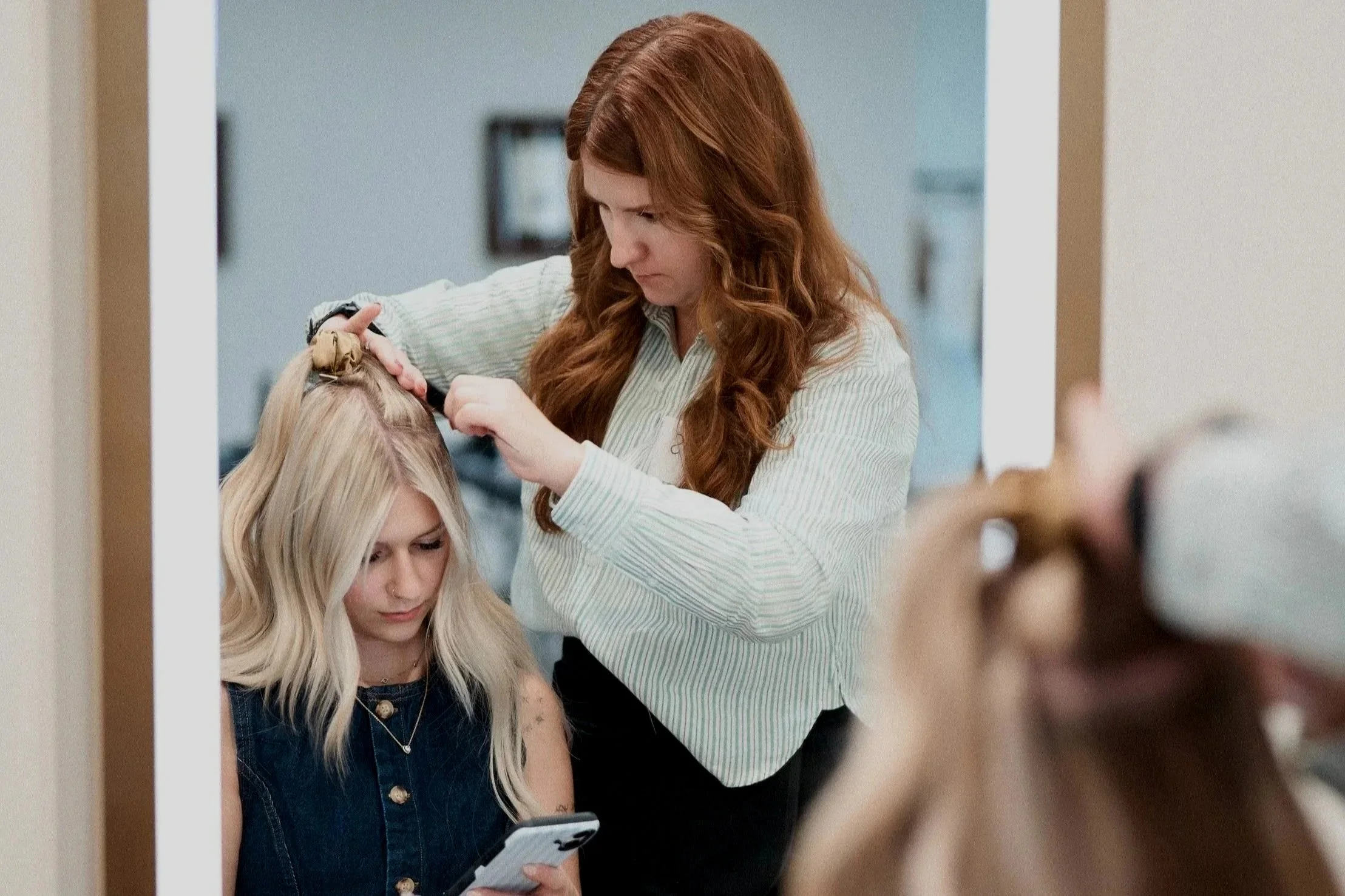 A hairdresser with long red hair styling a blonde woman's hair while she looks at her phone.