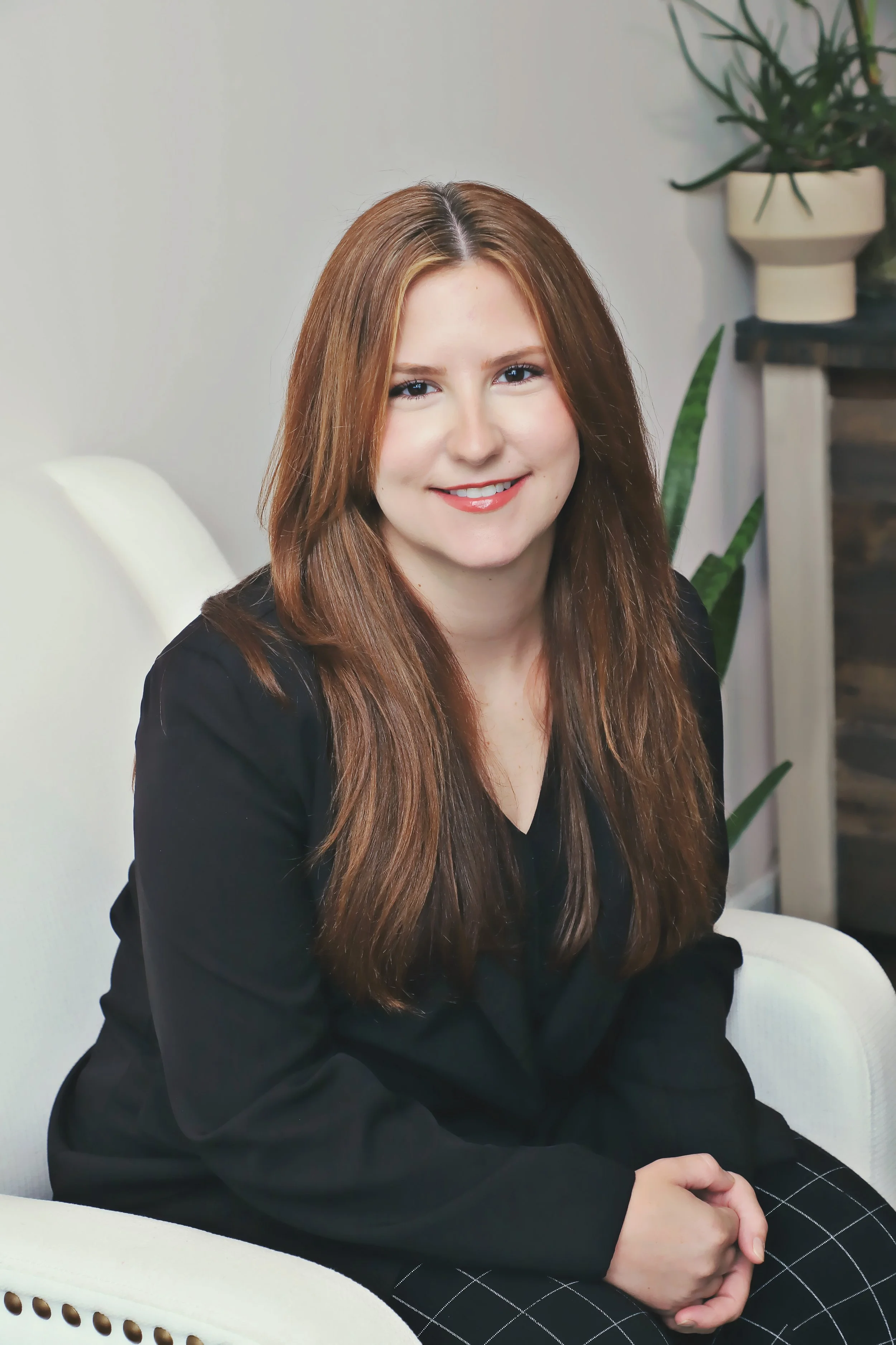 A woman with long, light brown hair and fair skin sitting on a white chair, smiling at the camera, wearing a black blazer and patterned pants, with indoor plants in the background.