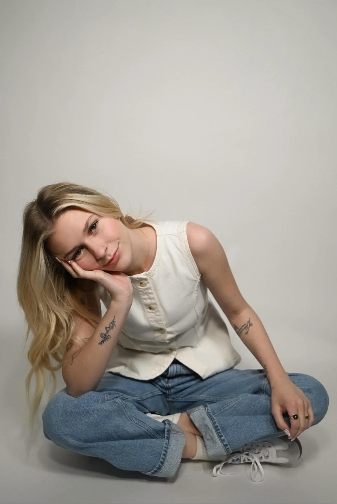 A young woman with long blonde hair, sitting cross-legged against a plain light-colored wall, resting her head on her hand, wearing a sleeveless white top, blue jeans, and white sneakers. Sophia Hromadko