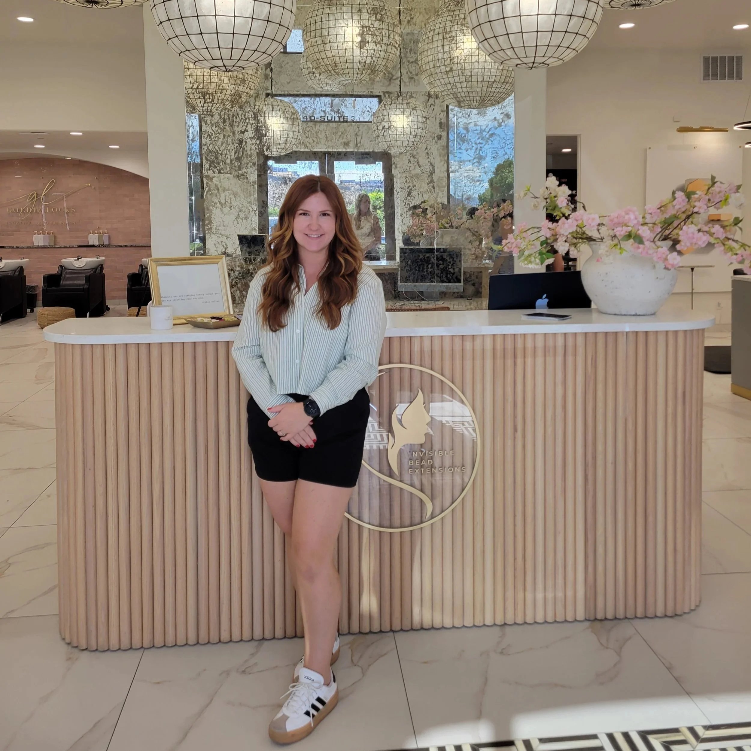 A woman with long wavy auburn hair and a striped long-sleeve shirt standing in front of a modern reception desk with a logo that reads 'Invisible Bead Extensions.' The desk is decorated with a large white vase of pink flowers. The background features a mirror, lounge chairs, and paper lanterns hanging from the ceiling.