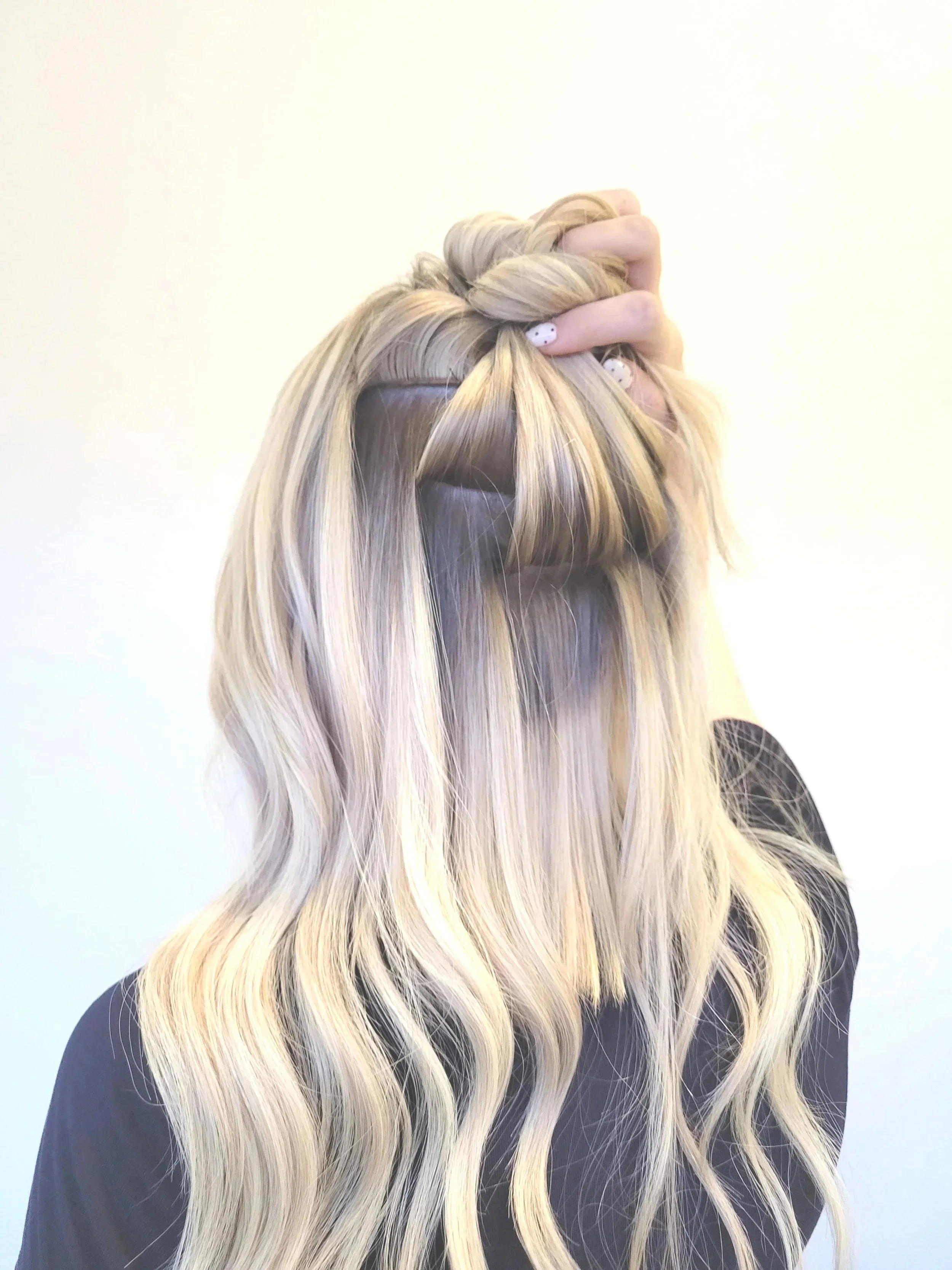 A woman with long, wavy blonde hair is holding her hair up, revealing dark roots. She has patterned nail art on her fingernails and is wearing a black top. The background is plain and light-colored.
