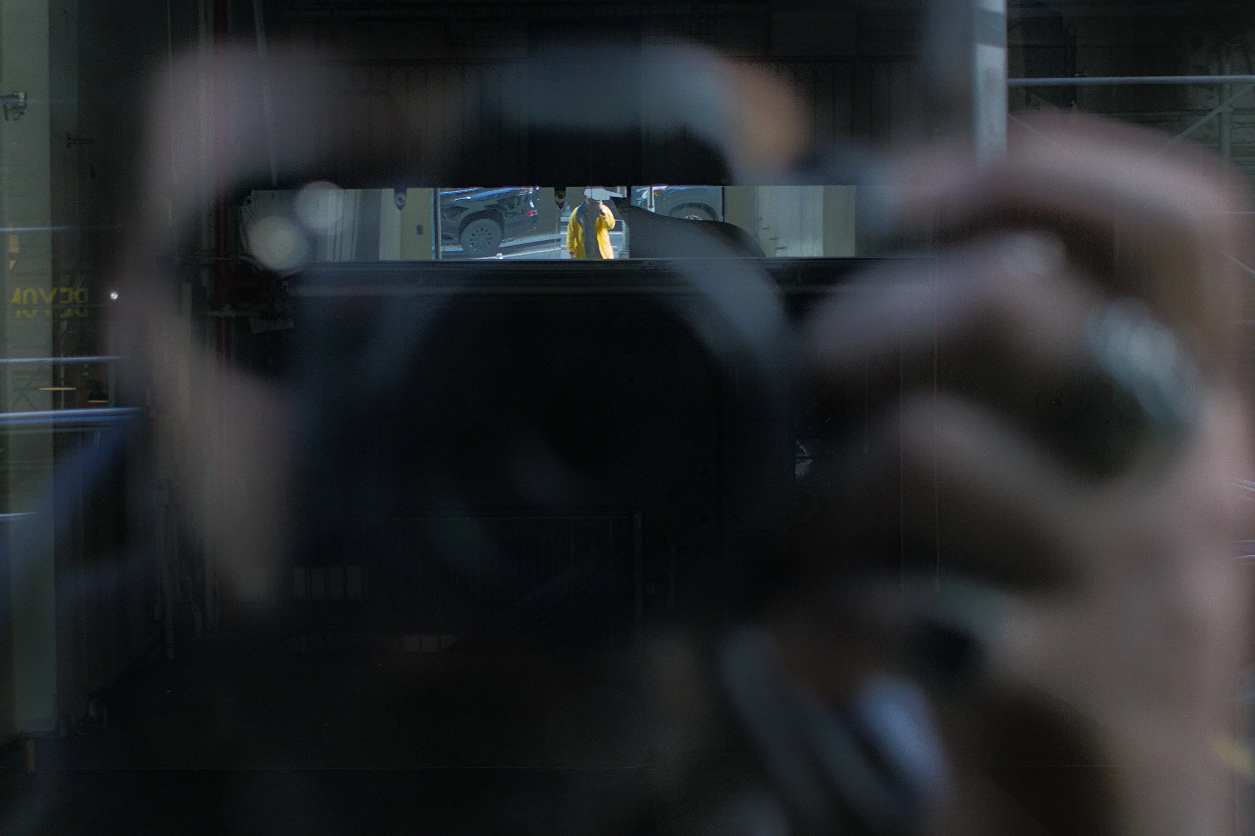 Photo taken through a glass window showing a person in a yellow jacket, holding a phone or camera, standing outside in a parking lot with cars visible. The scene is blurry with some reflections and a person's hand visible in the foreground.