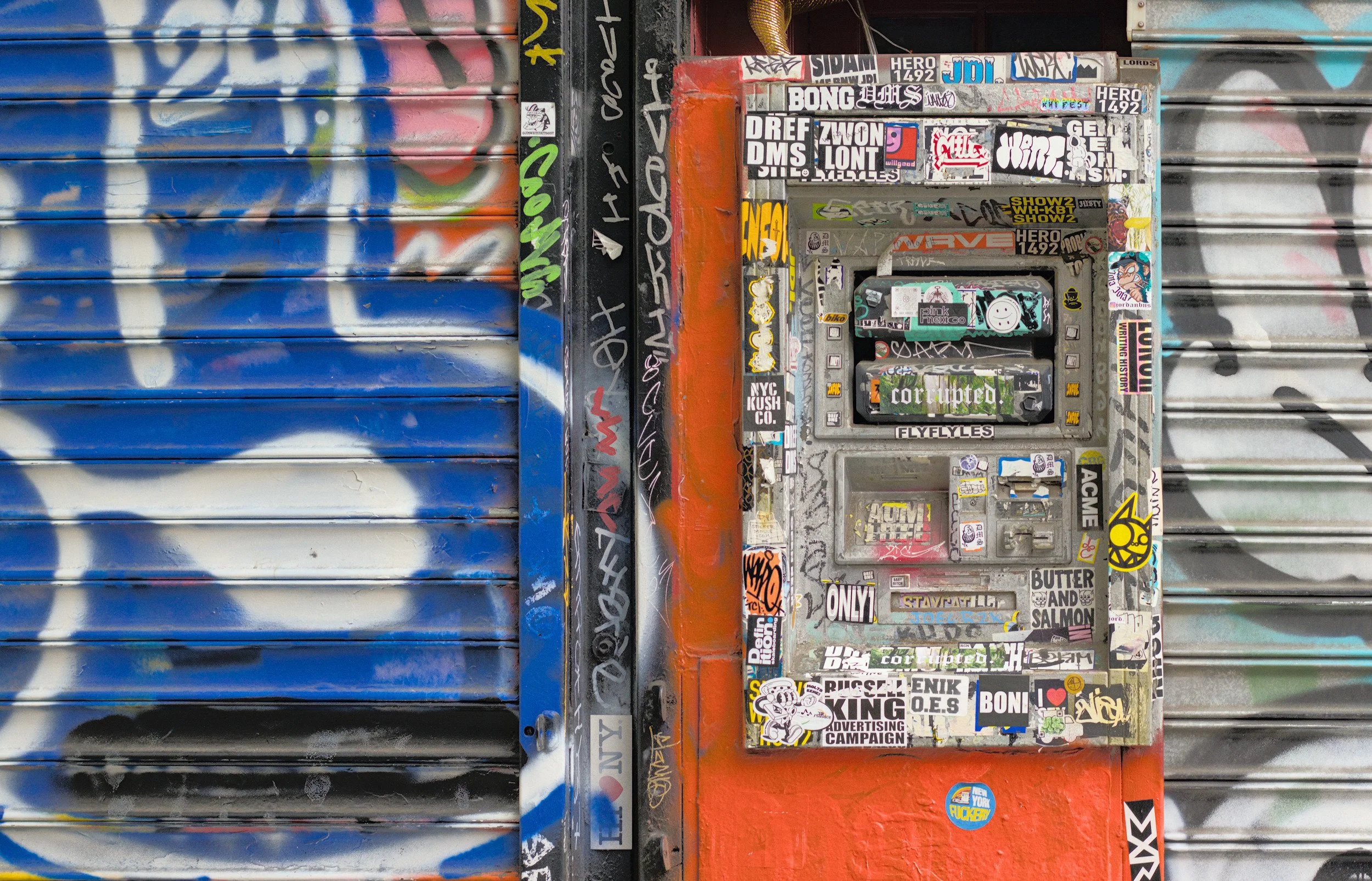 ATM machine covered in graffiti stickers and tags on city street. Gritty urban detail photography by Derek Ronald.