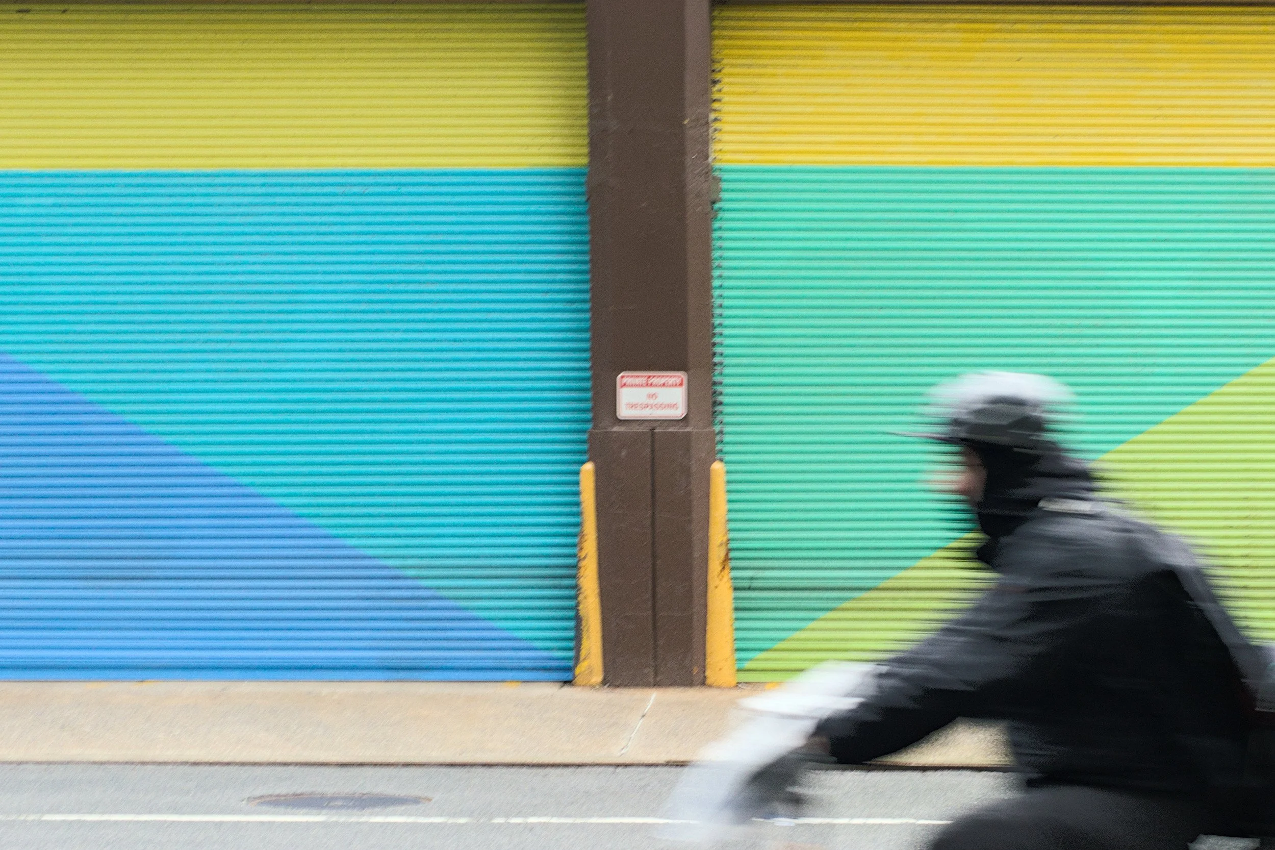 Motion blur of cyclist passing colorful teal and yellow striped wall. Urban street photography by Derek Ronald.