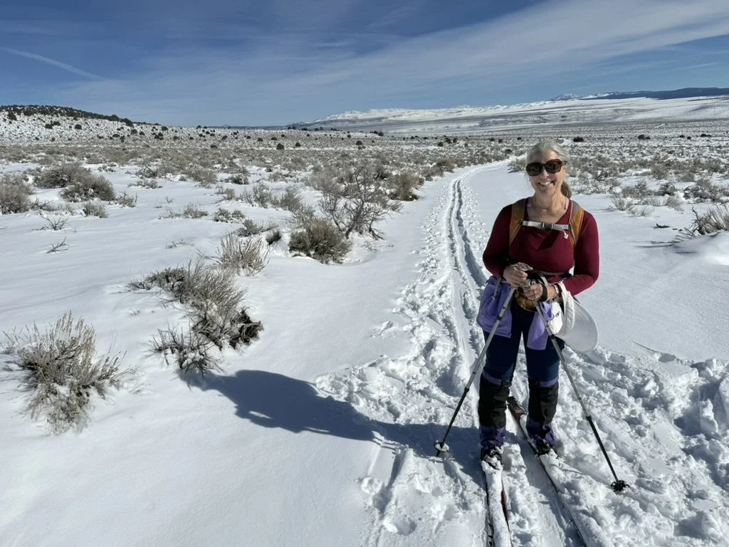 A smiling woman wearing sunglasses and winter clothing, holding ski poles, stands on a snow-covered trail in a vast, open winter landscape with sparse bushes and mountains in the background.
