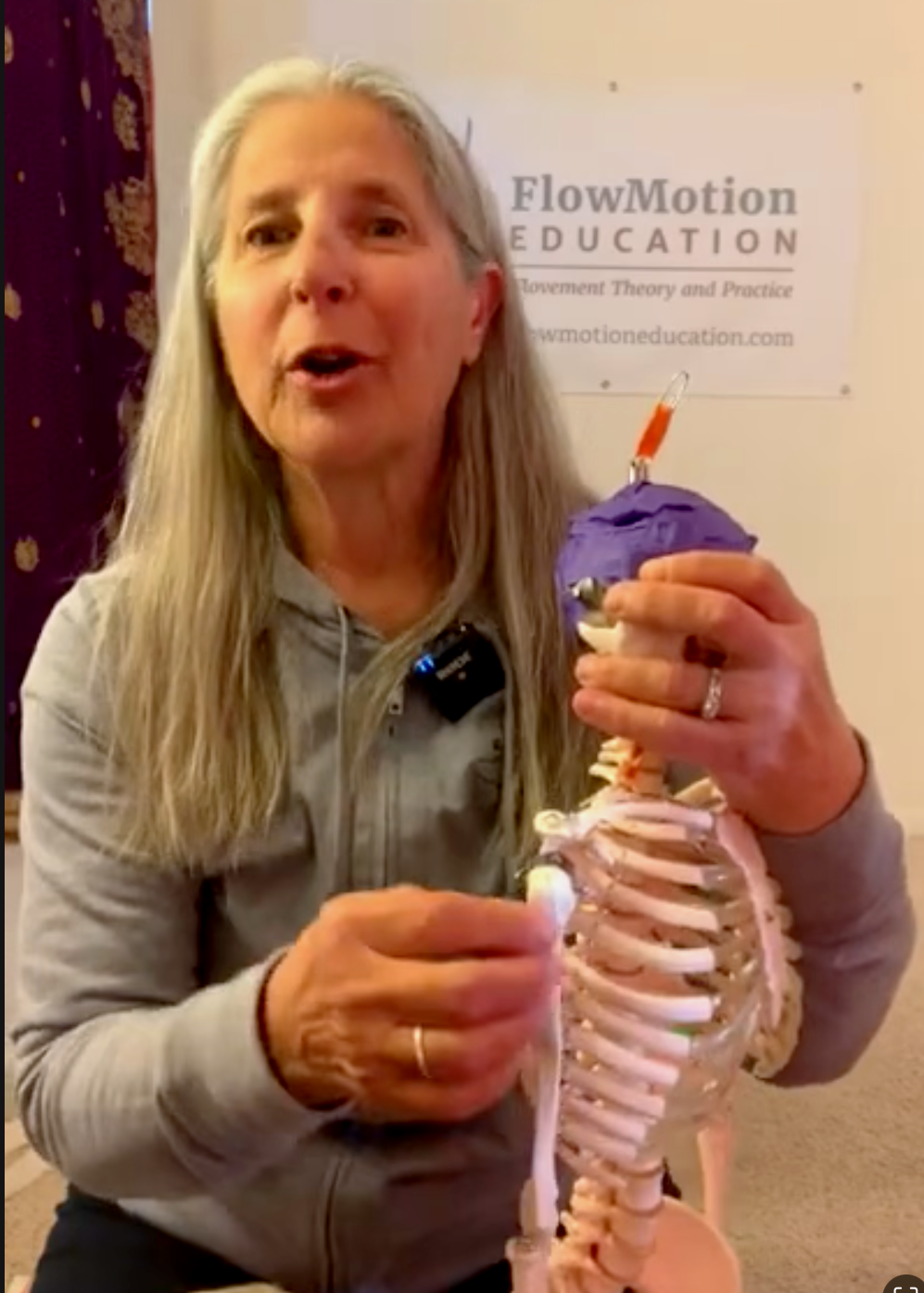 A woman with long gray hair holding a model of a human skeleton with a purple brain and orange needle, standing in front of a sign that reads 'FlowMotion Education' at an educational event.