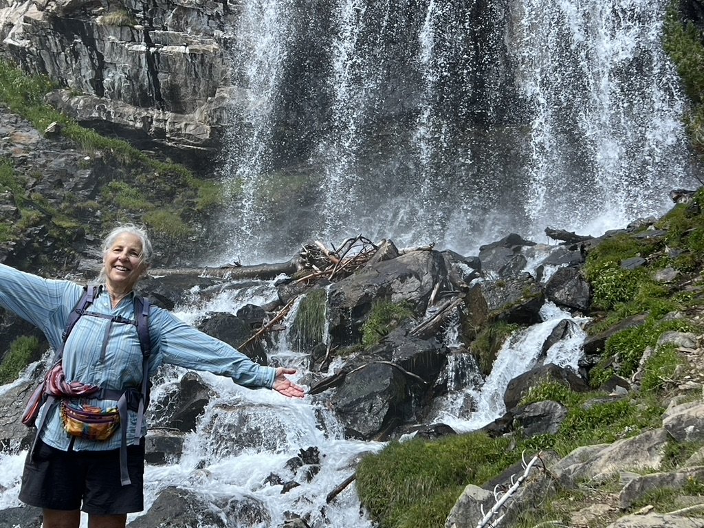 An elderly woman smiling with outstretched arms standing in front of a waterfall flowing over rocks and greenery on a mountain trail.