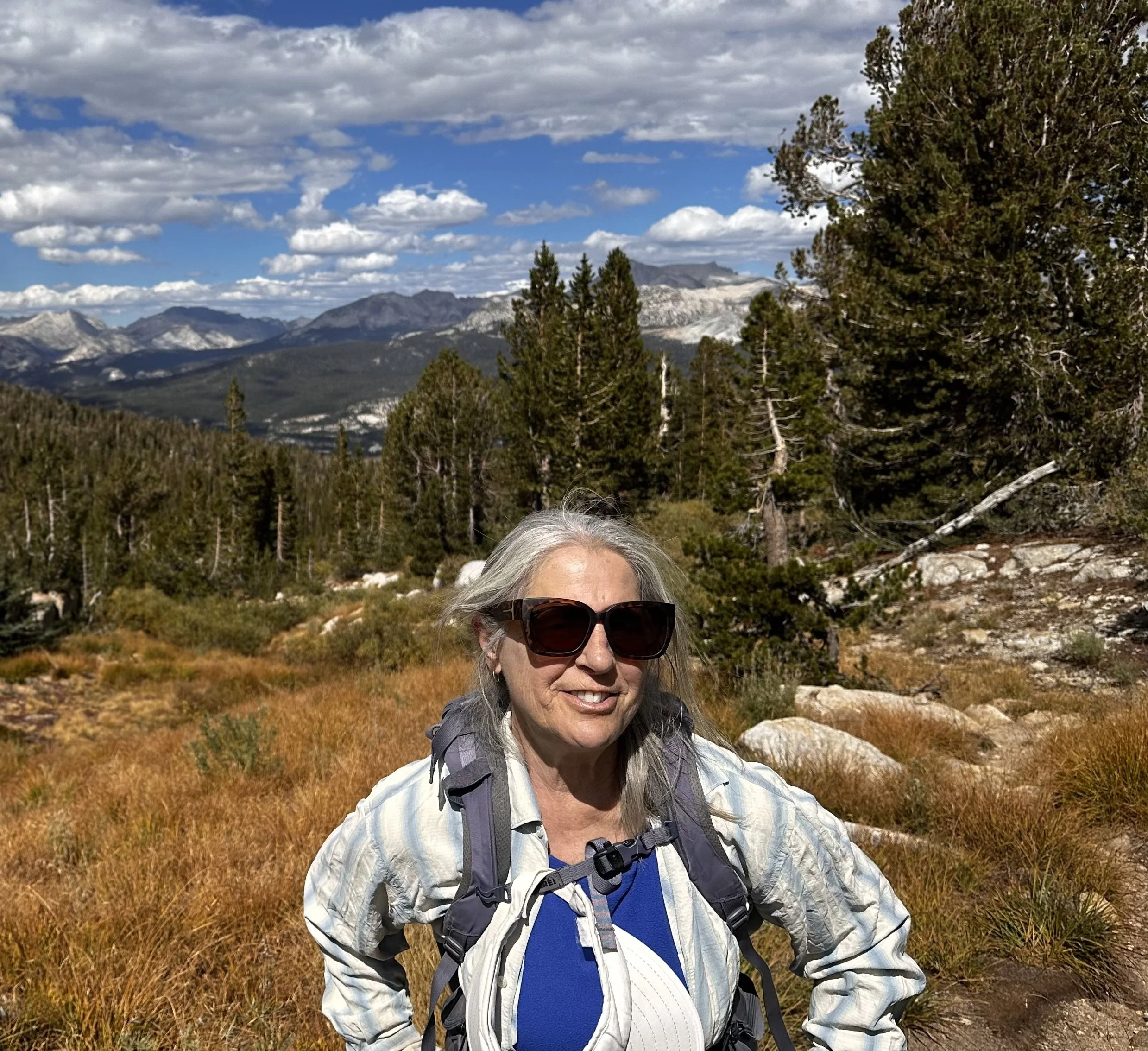 A woman with gray hair wearing sunglasses and a plaid shirt smiles in a mountain landscape with trees, mountains, and a partly cloudy sky.